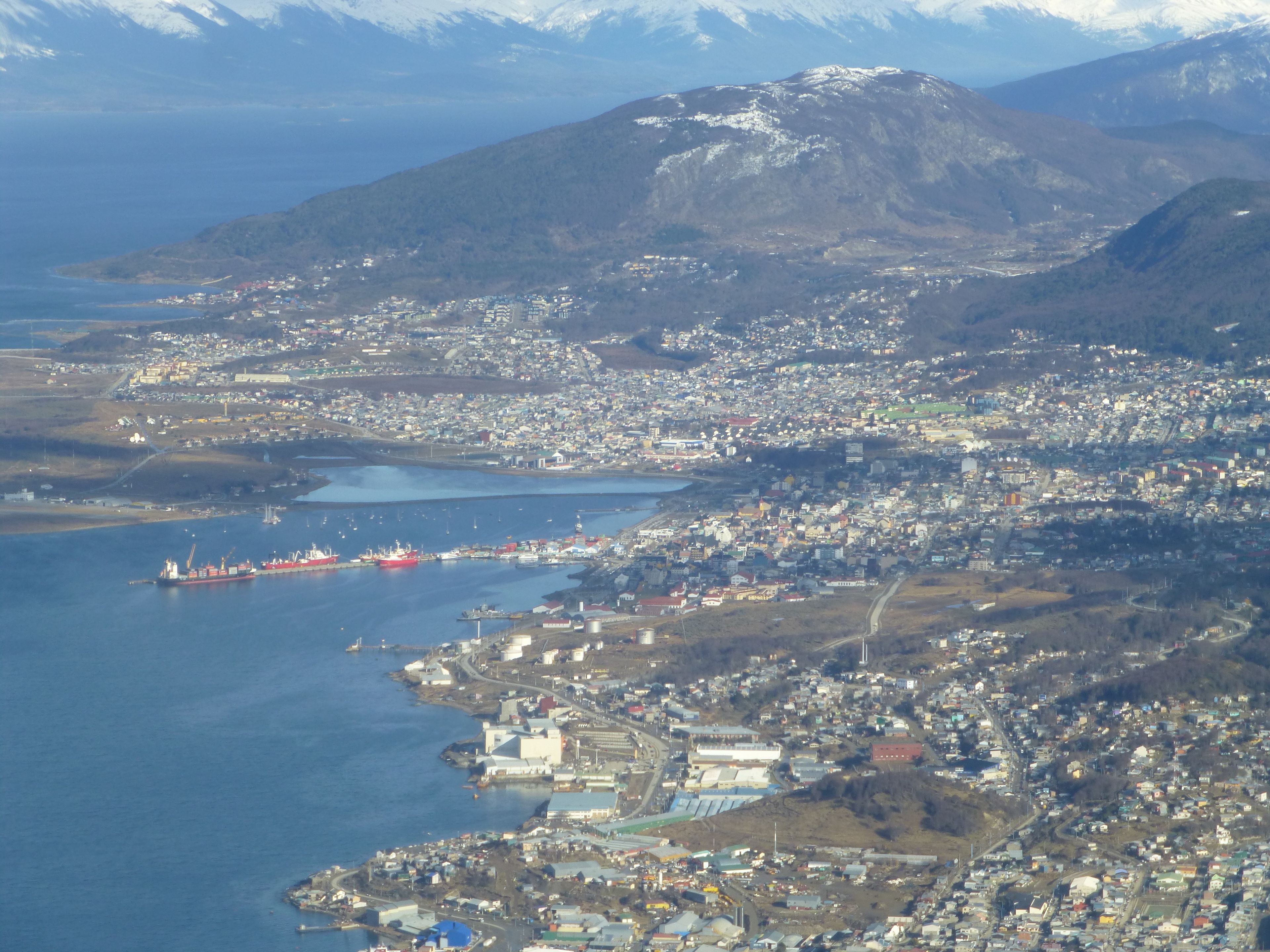 Vista aérea de un pueblo costero con un puerto, edificios y una montaña al fondo.