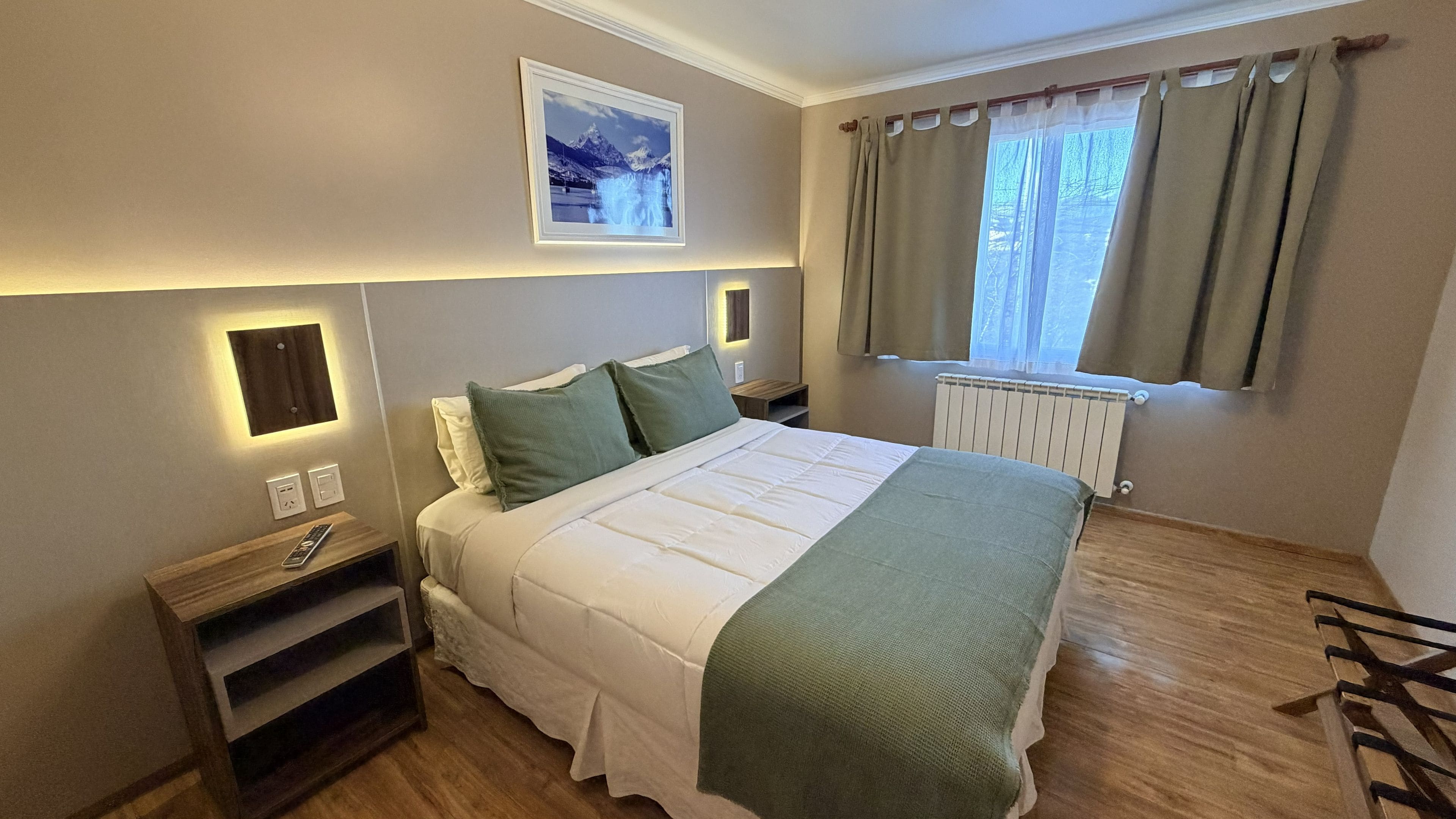 Habitación de hotel con cama, mesitas de noche, radiador, ventana con cortinas y un cuadro colgado en la pared.
