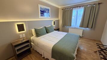 Habitación de hotel con cama, mesitas de noche, radiador, ventana con cortinas y un cuadro colgado en la pared.