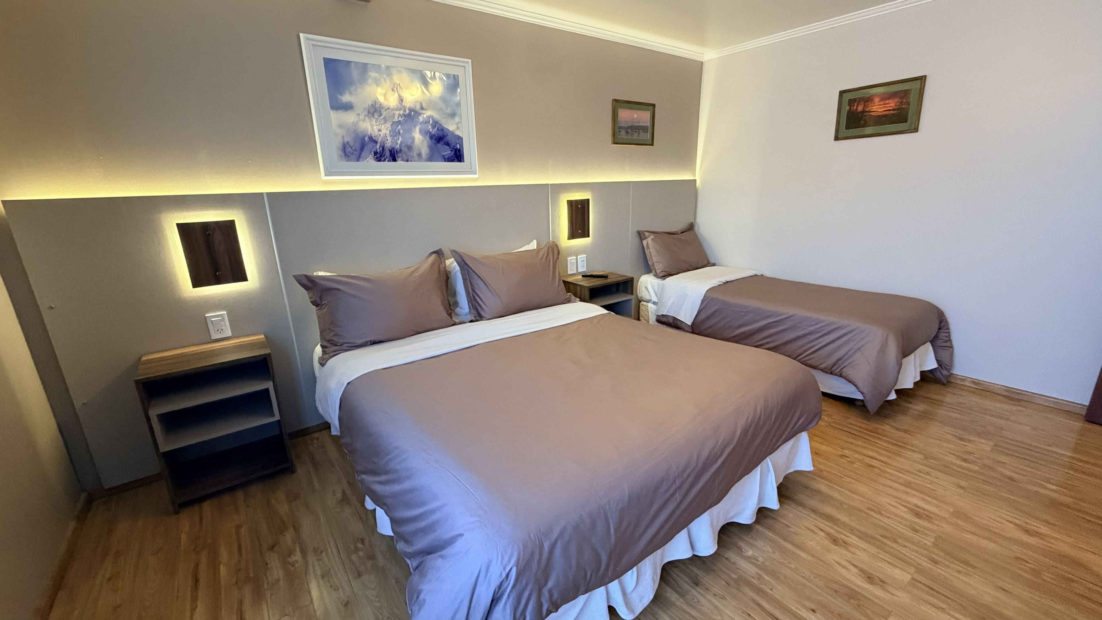 A hotel room with two beds, a nightstand, a wall-mounted lamp, and a framed picture.