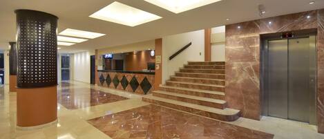A modern lobby with a marble floor, a reception desk, a staircase, and an elevator.
