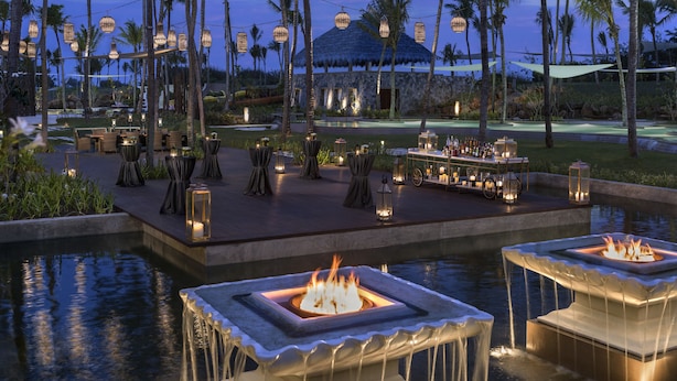 Zona de piscina en un resort, con fogones, mobiliario exterior y palmeras.