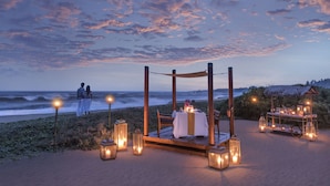 Un montaje para comer en la playa con una mesa y sillas bajo una pérgola, rodeado de faroles.