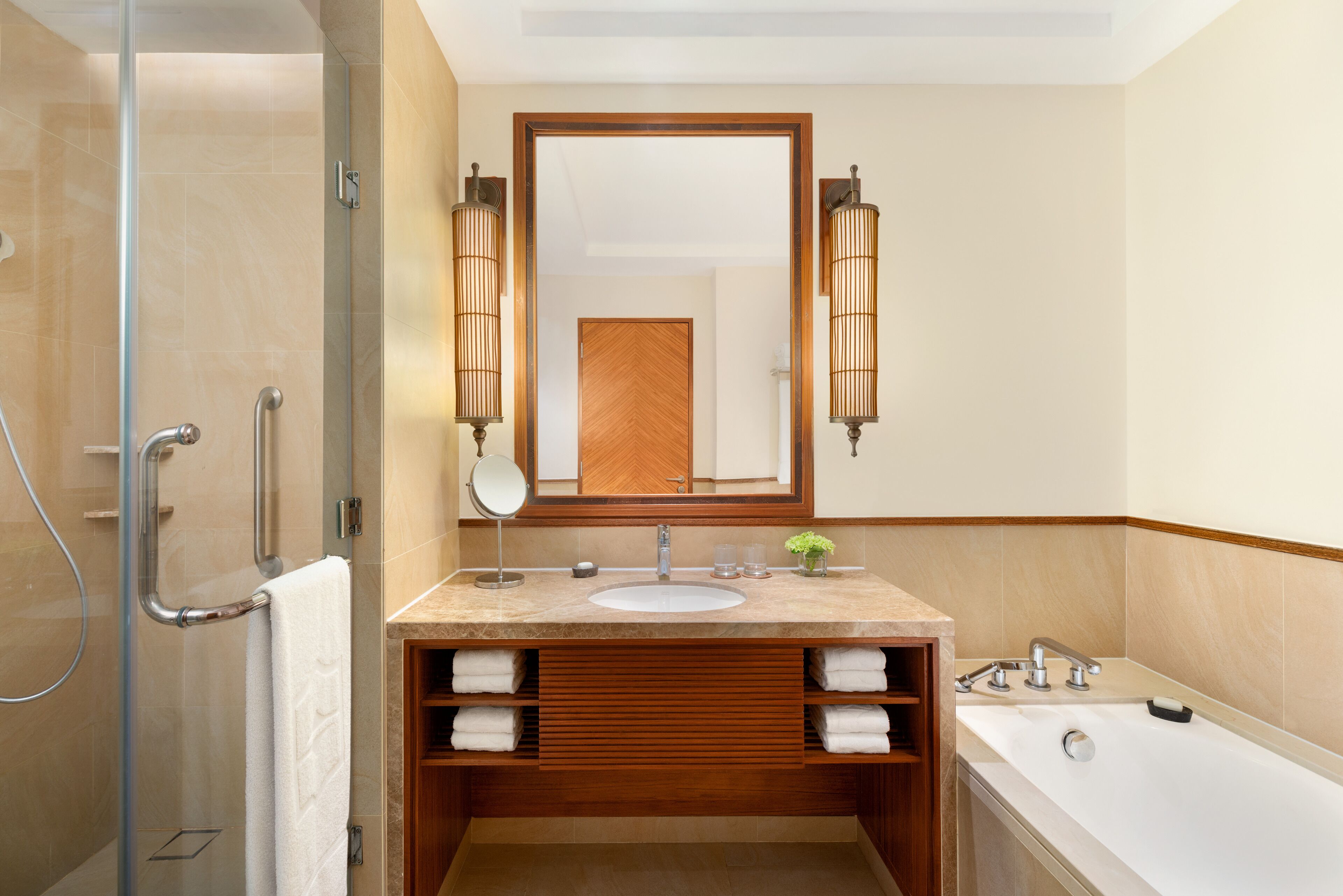 A modern bathroom with a glass shower enclosure, a wooden vanity with a sink, and a large mirror with wall sconces.