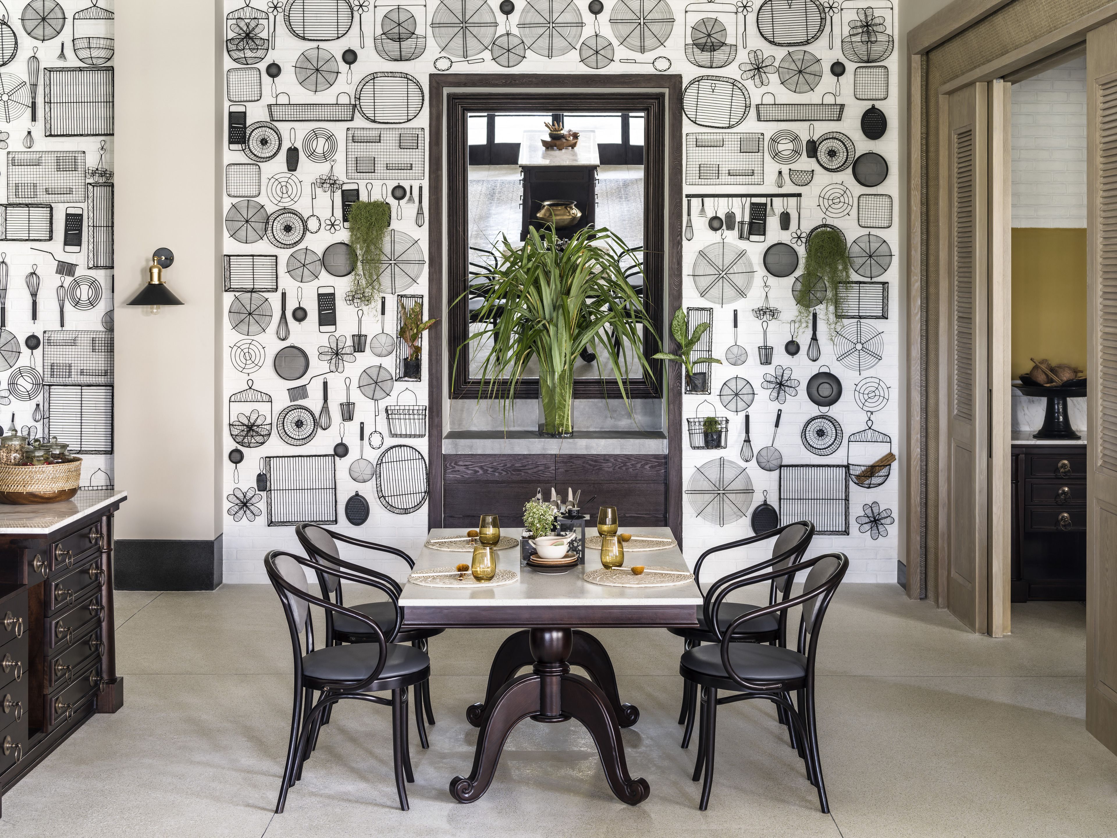 A dining area with a table set for two, surrounded by black chairs. A mirror with a dark frame is centered above the table, reflecting a plant and a window. The walls are decorated with a pattern of kitchen utensils and cookware.
