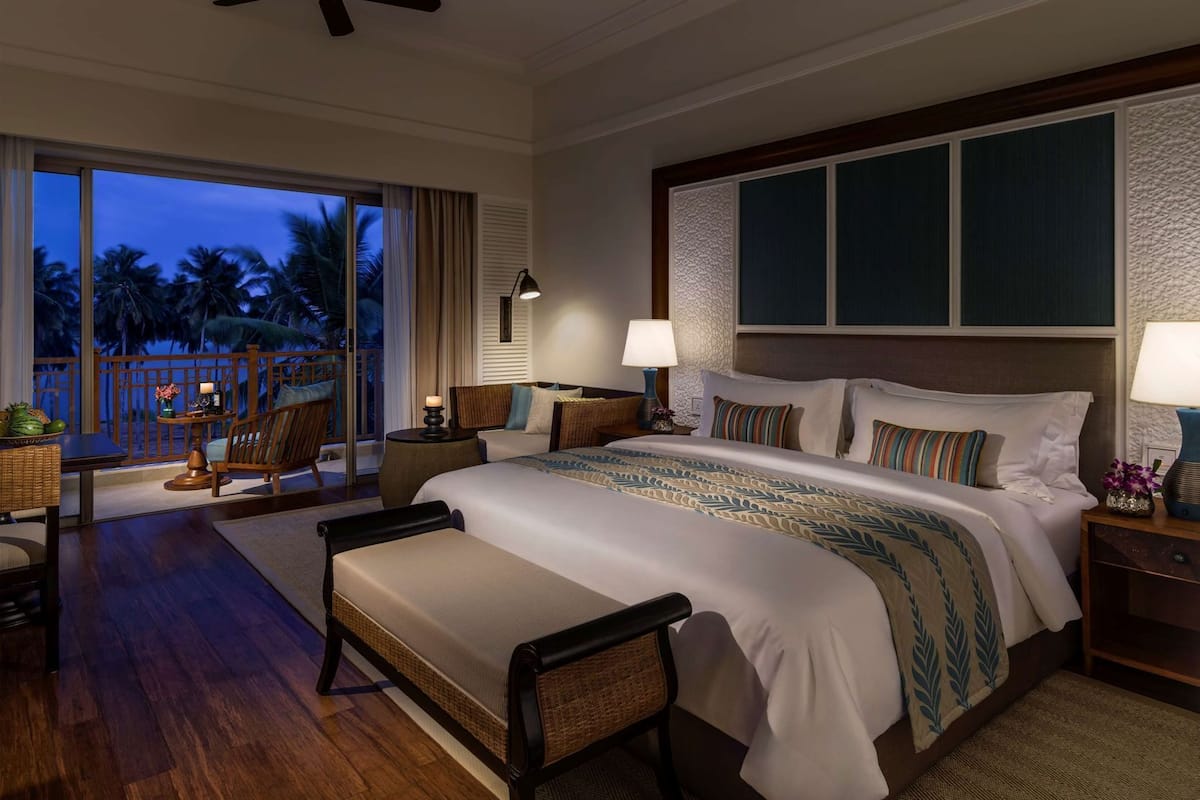 Een hotelkamer met een groot bed, een balkon met uitzicht op palmbomen, een zithoek met een tafel en stoelen, en een plafondventilator.