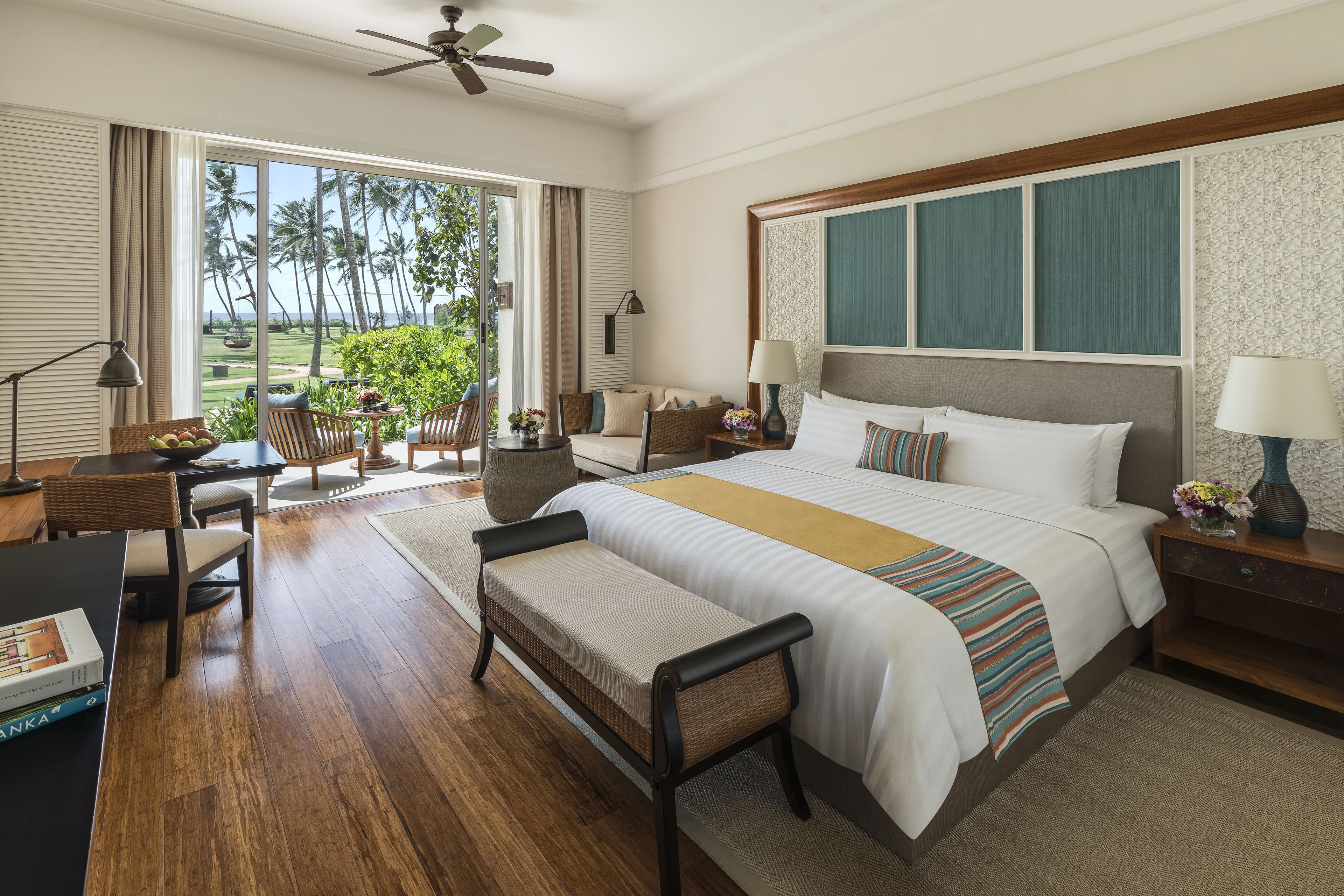 A hotel room with a large bed, a desk with a chair, a bench, and a view of palm trees through the window.