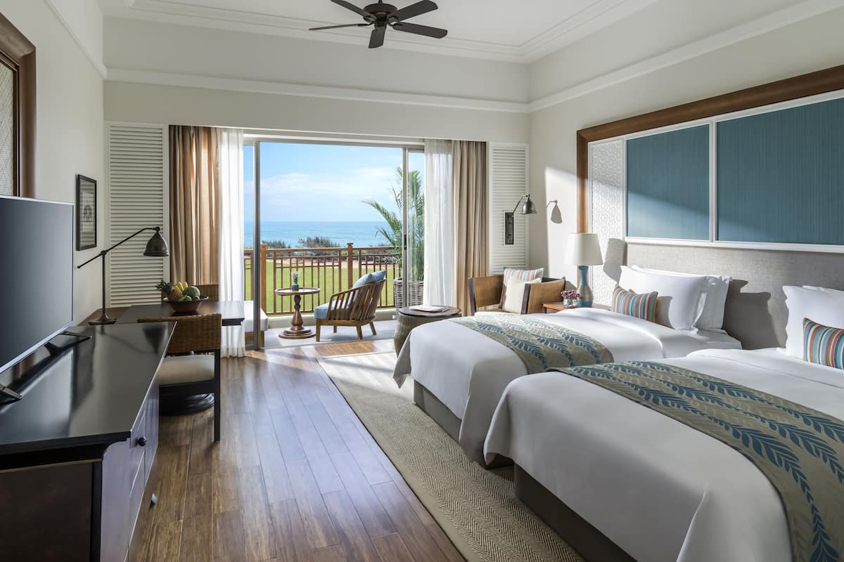 Hotelkamer met twee bedden, een televisie, een bureau, een balkon met uitzicht op zee en een plafondventilator.