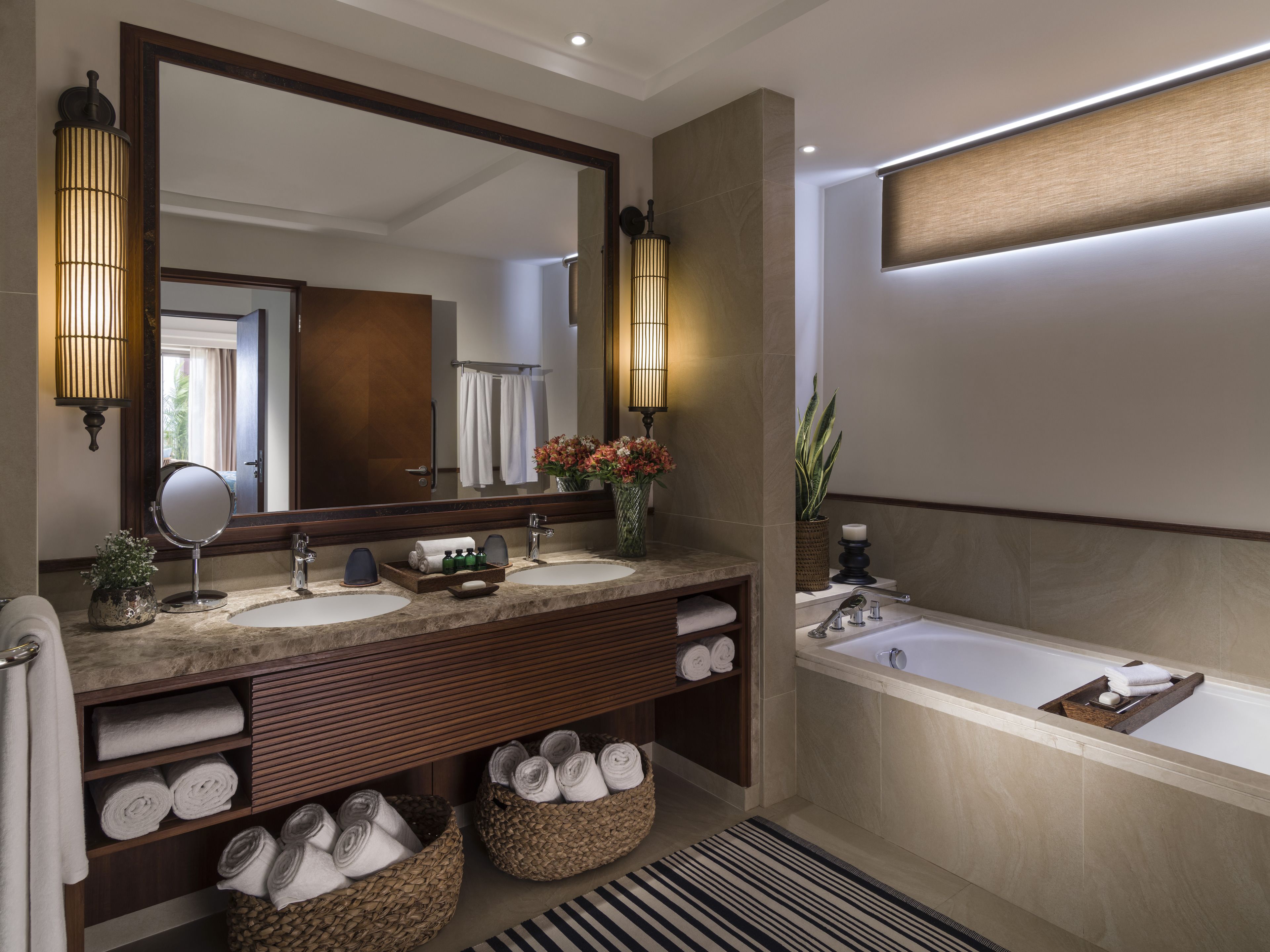 A modern bathroom with two sinks, a large mirror, and a bathtub. There are towels and toiletries on the counter, and a potted plant on the side.