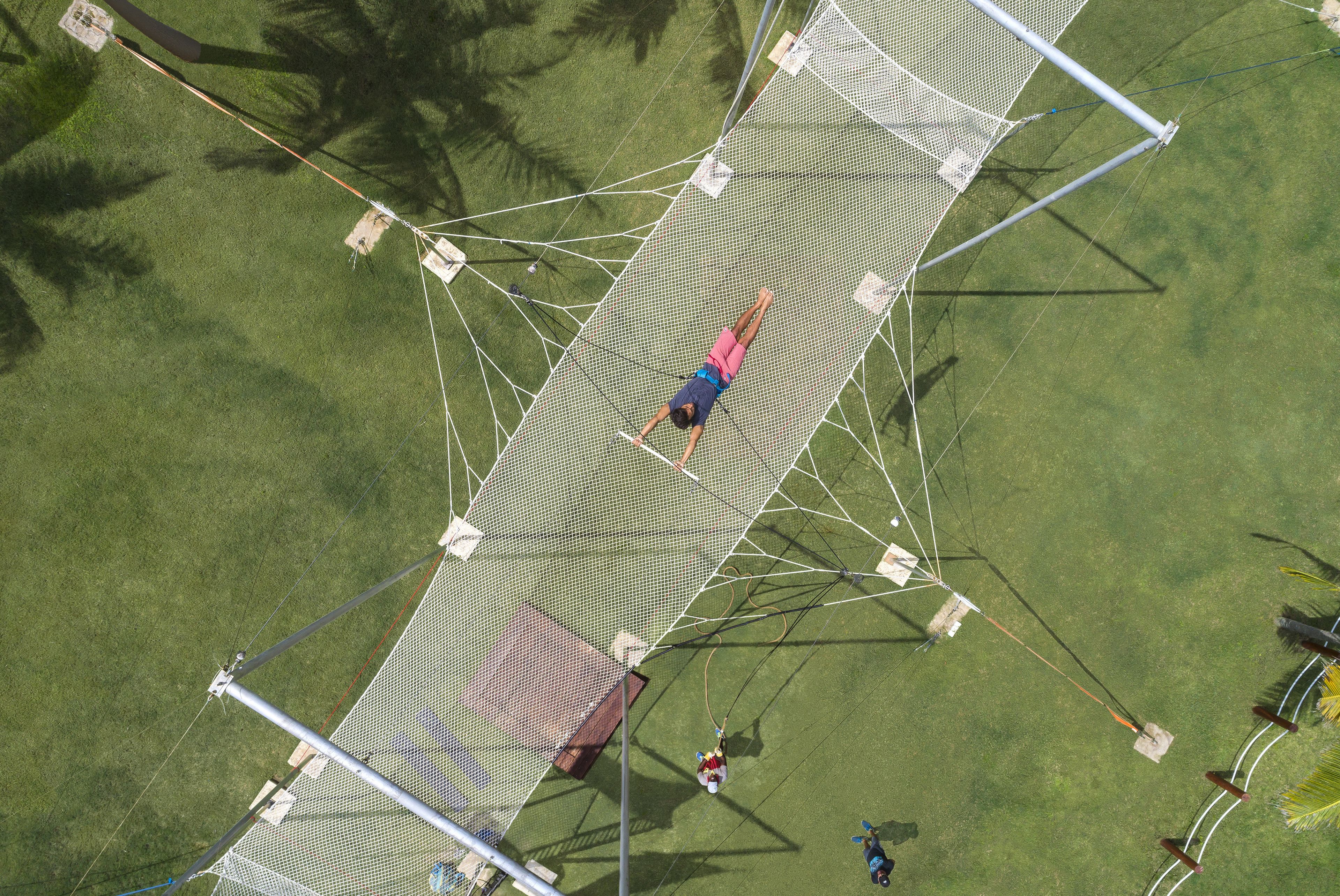 An aerial view of a netted zip line structure on a grassy field with trees in the background.