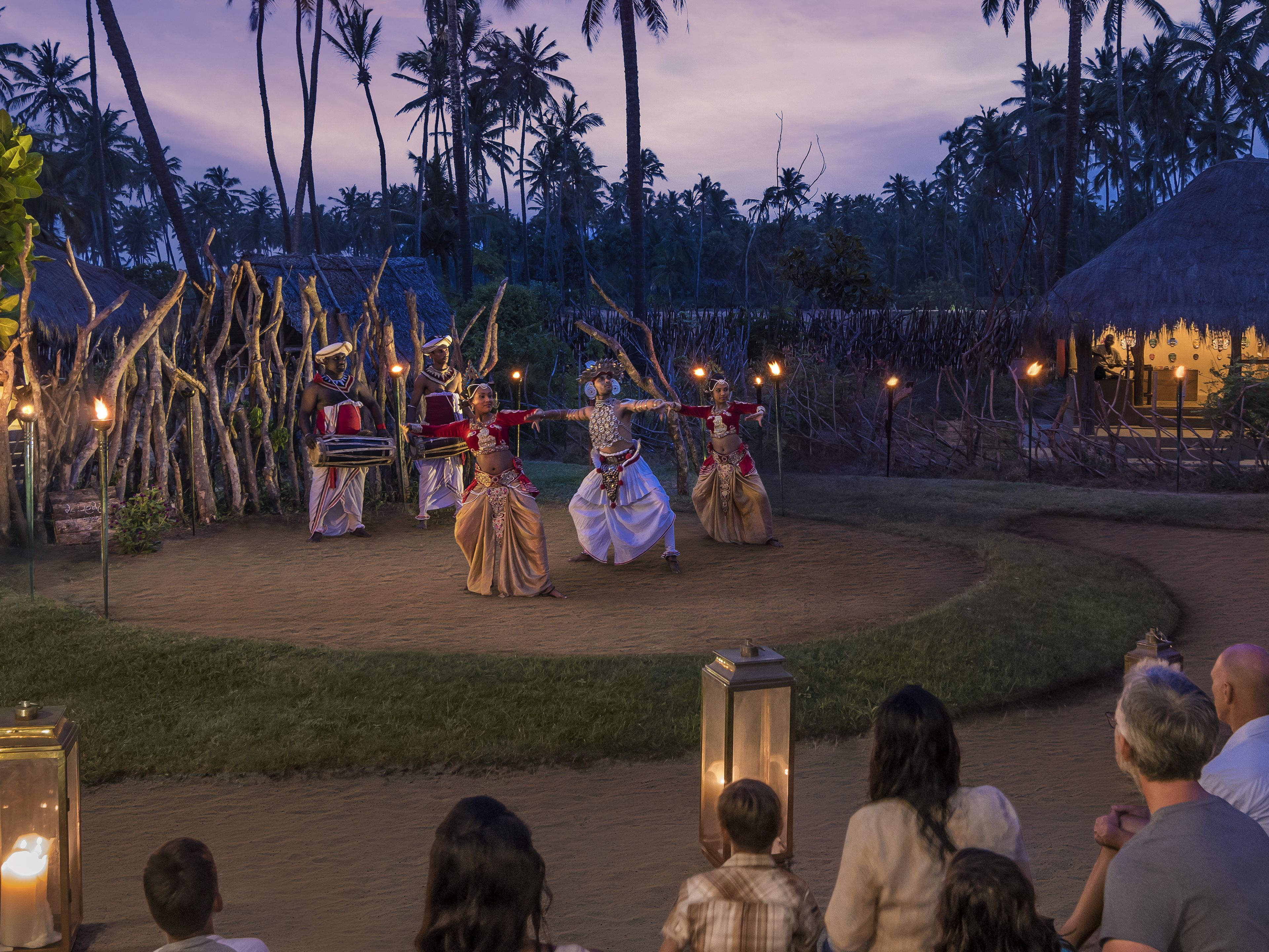 A traditional dance performance in an outdoor setting with a dirt path, palm trees, and a thatched-roof structure in the background.