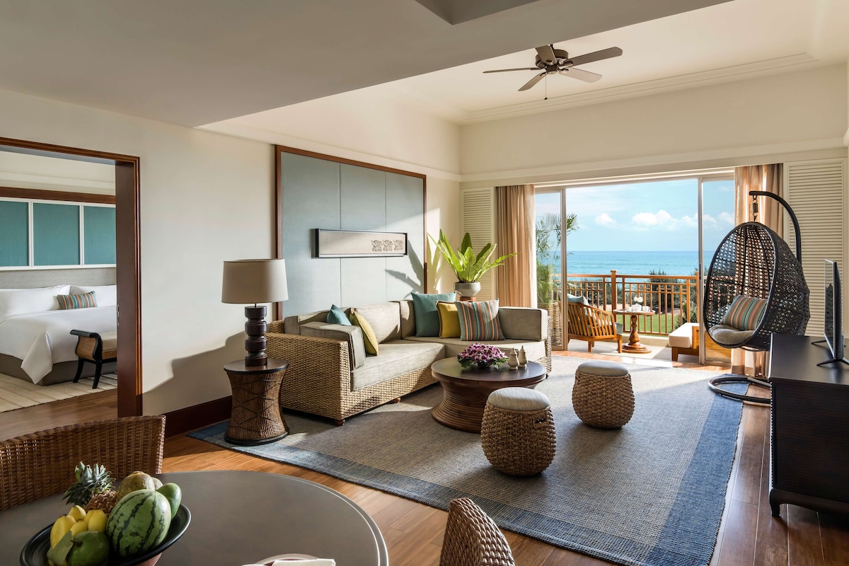 Une chambre d’hôtel moderne avec un balcon, un canapé, une table à manger avec des fruits et un fauteuil suspendu.