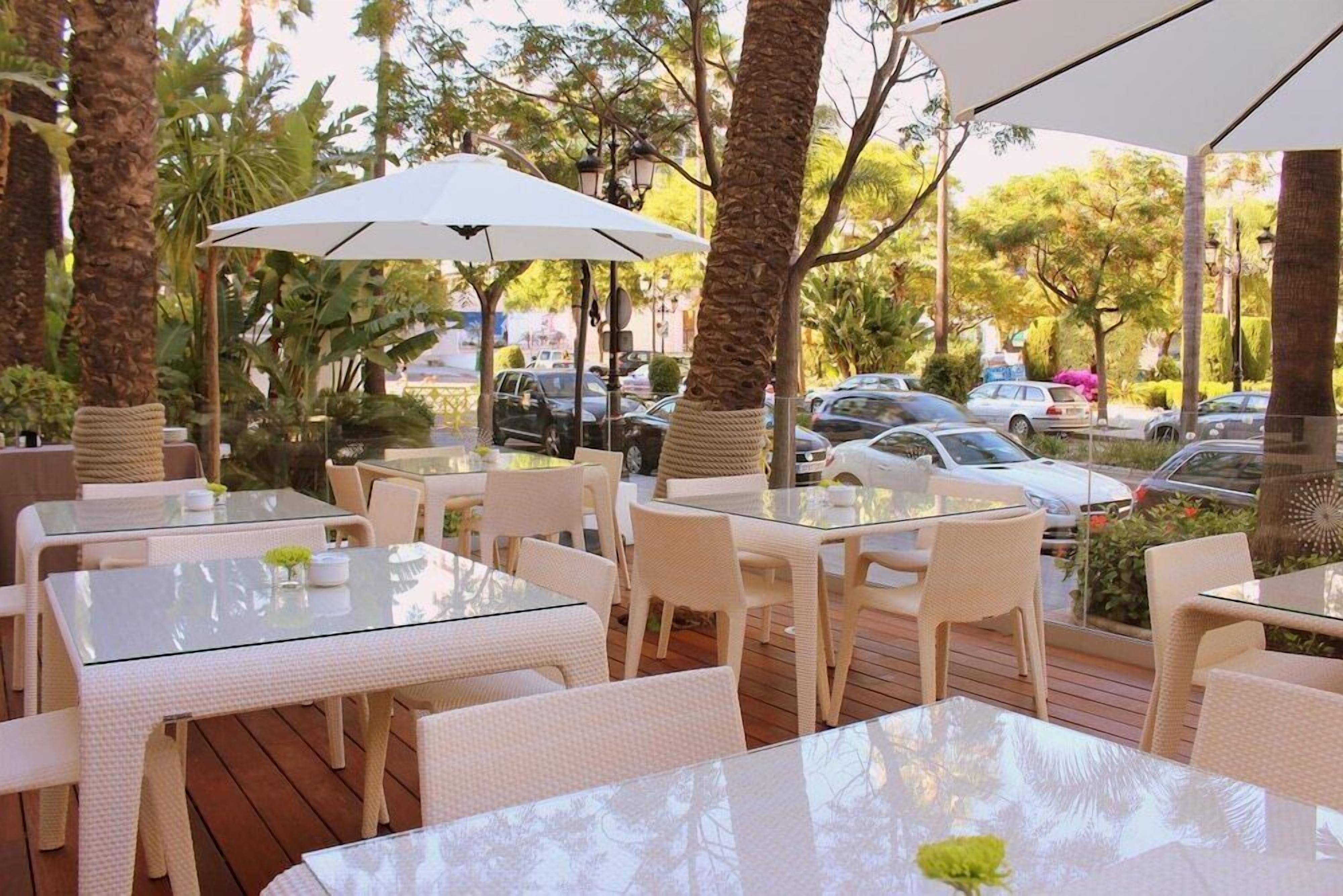 An outdoor patio area with glass-top tables and white wicker chairs, surrounded by palm trees and parked cars.