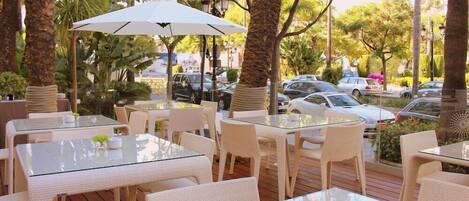An outdoor patio area with glass-top tables and white wicker chairs, surrounded by palm trees and parked cars.