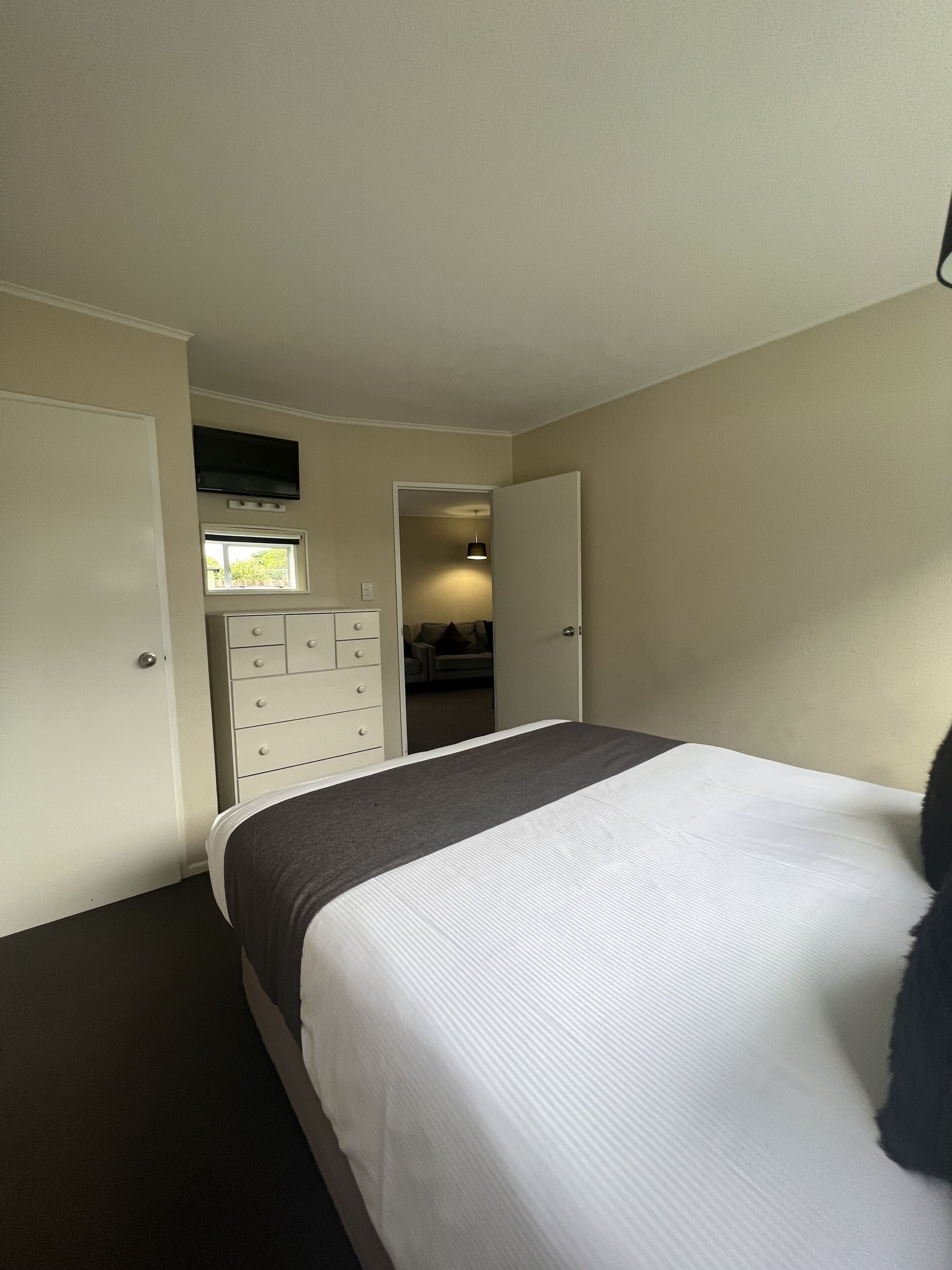 A bedroom with a bed, a dresser, a TV mounted above a window, and a door leading to another room.