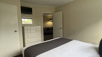 Un dormitorio con una cama, un aparador, un televisor sobre una ventana y una puerta que da a otra habitación.