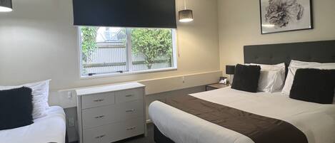 Habitación de hotel con dos camas, un escritorio, una silla, una ventana con persianas, una lámpara de pared y un cuadro enmarcado.