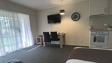Een moderne hotelkamer met een groot bed, een bureau met twee stoelen, een flatscreen televisie aan de muur, een klok en een kitchenette met oven en magnetron.