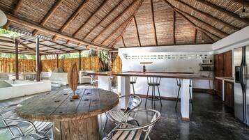 An outdoor dining area with a wooden table and metal chairs, a bar area with stools, and a kitchen with appliances.