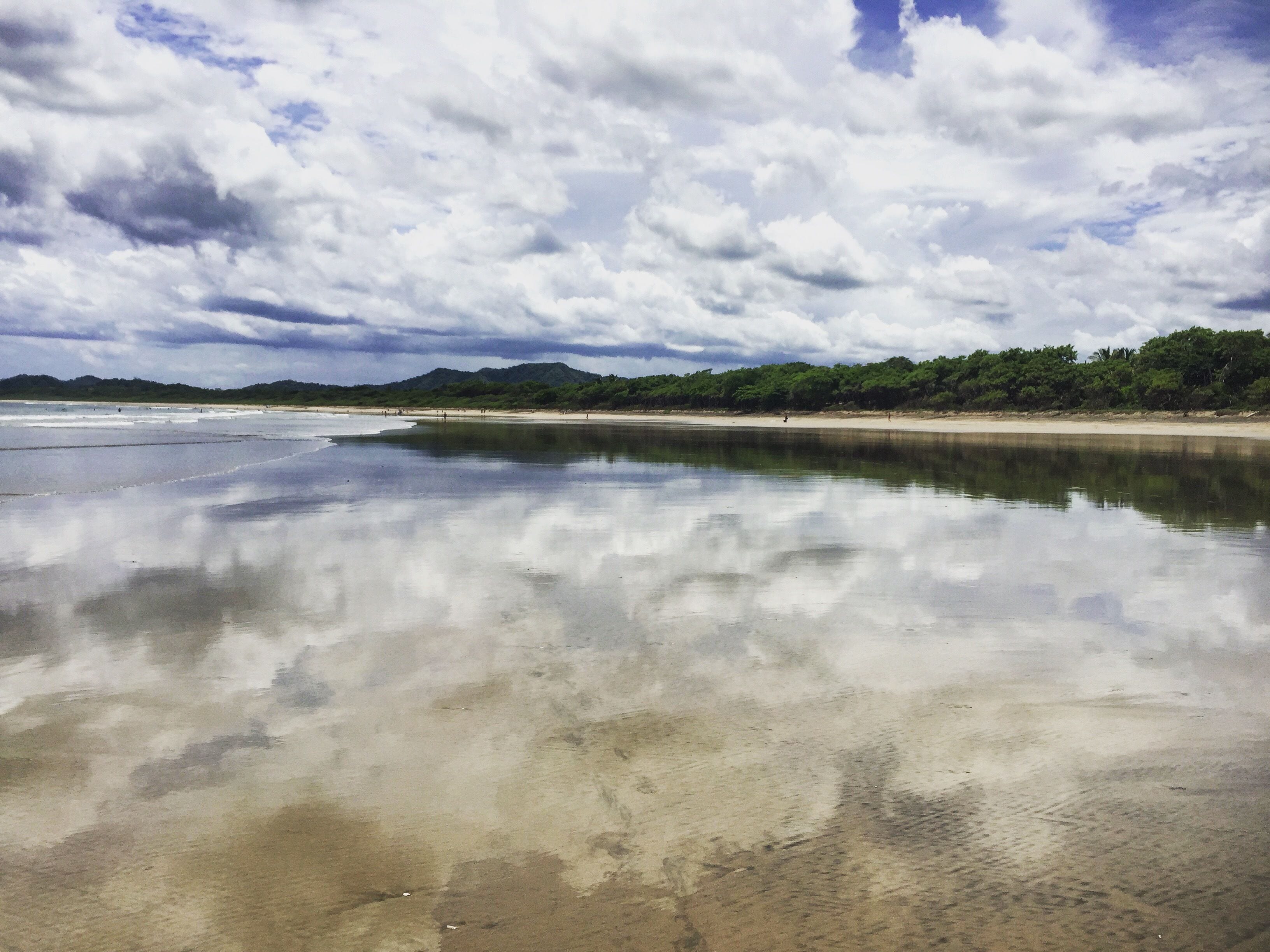 Una playa con aguas tranquilas que reflejan el cielo y colinas lejanas.