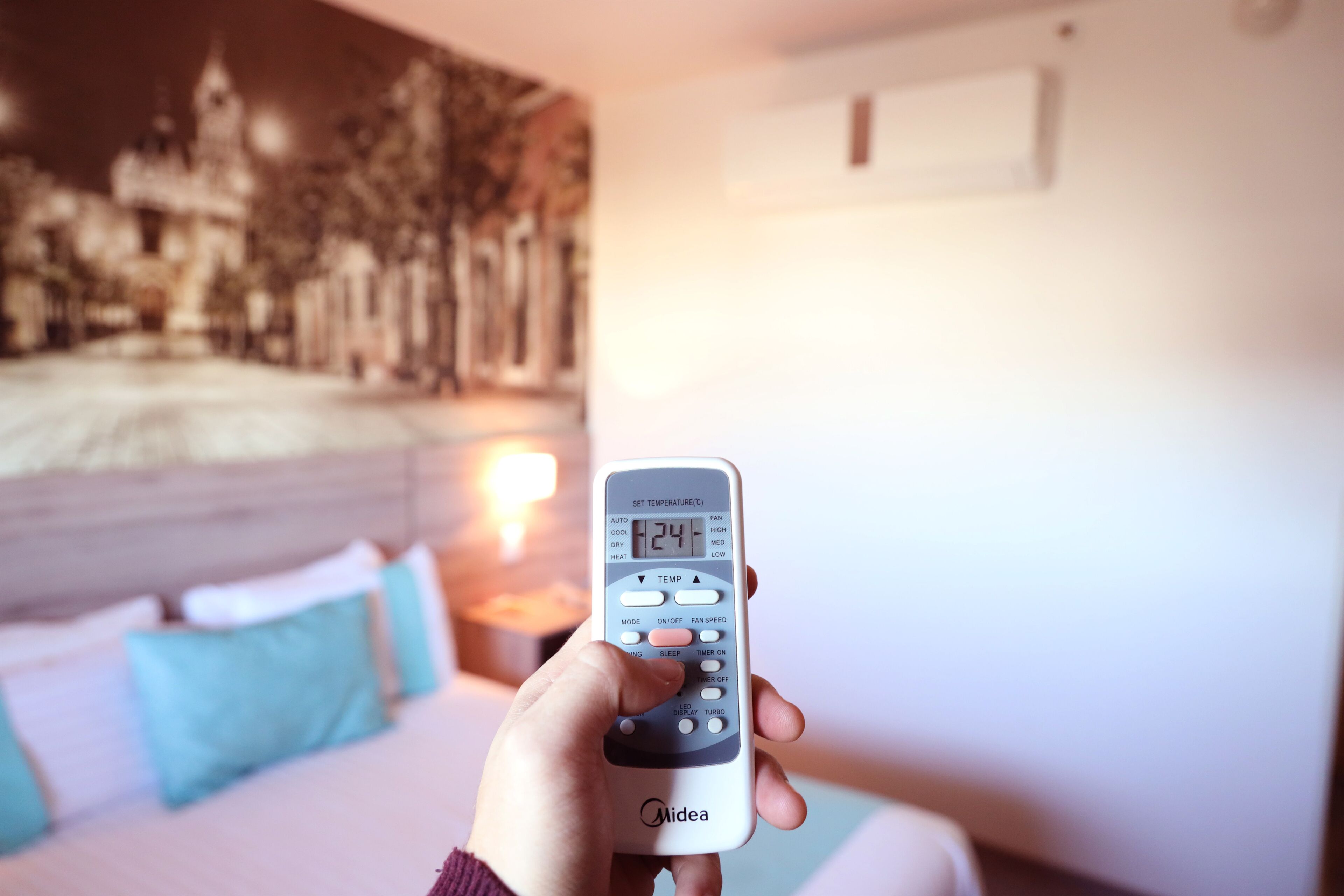 A hand holding a remote control with a digital display, a bed with pillows, and a wall with a painting.