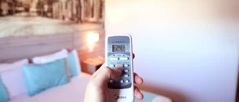 A hand holding a remote control with a digital display, a bed with pillows, and a wall with a painting.