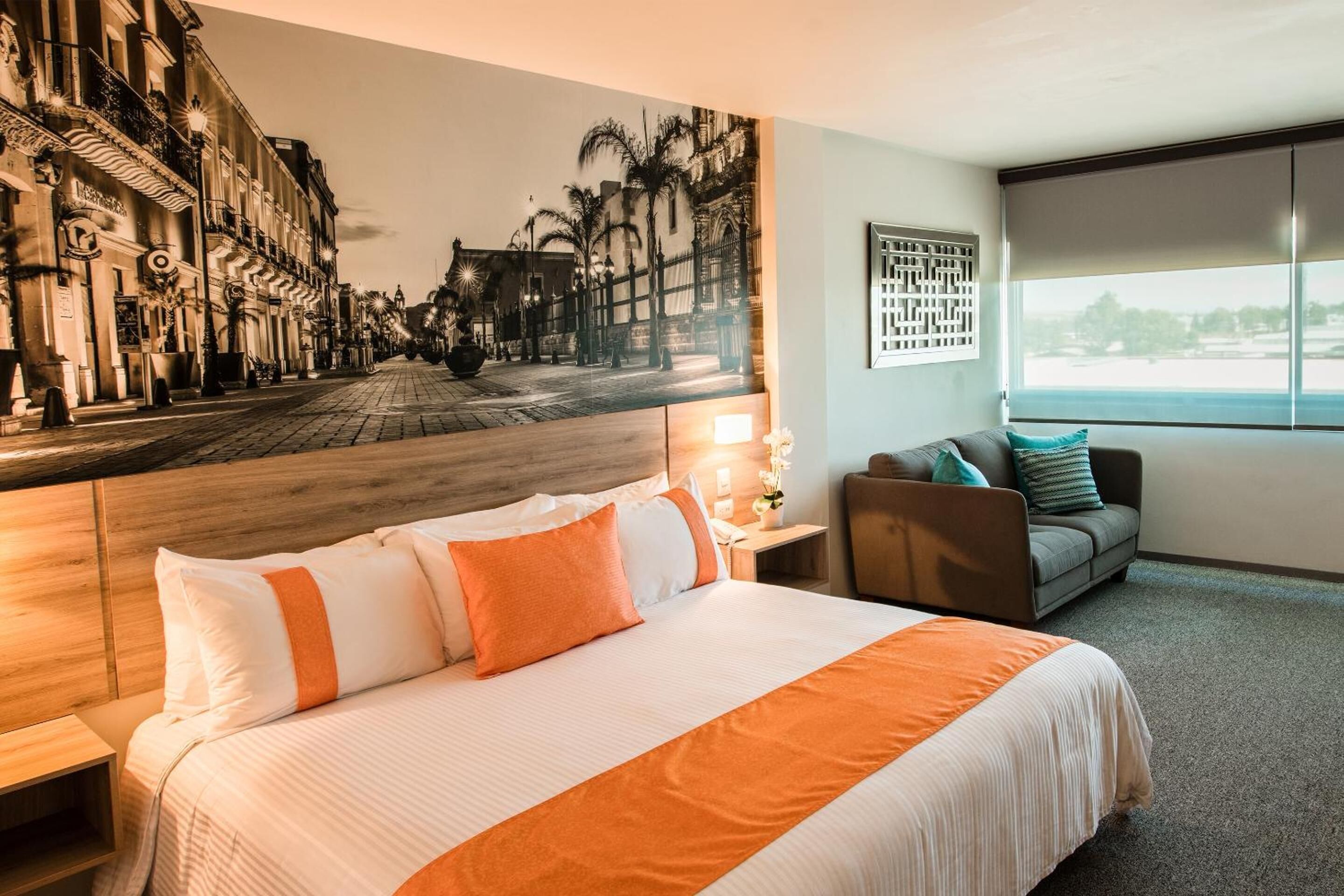 A hotel room with a large bed, a view of a cityscape through a window, a seating area with a sofa and armchairs, and a wall-mounted artwork.
