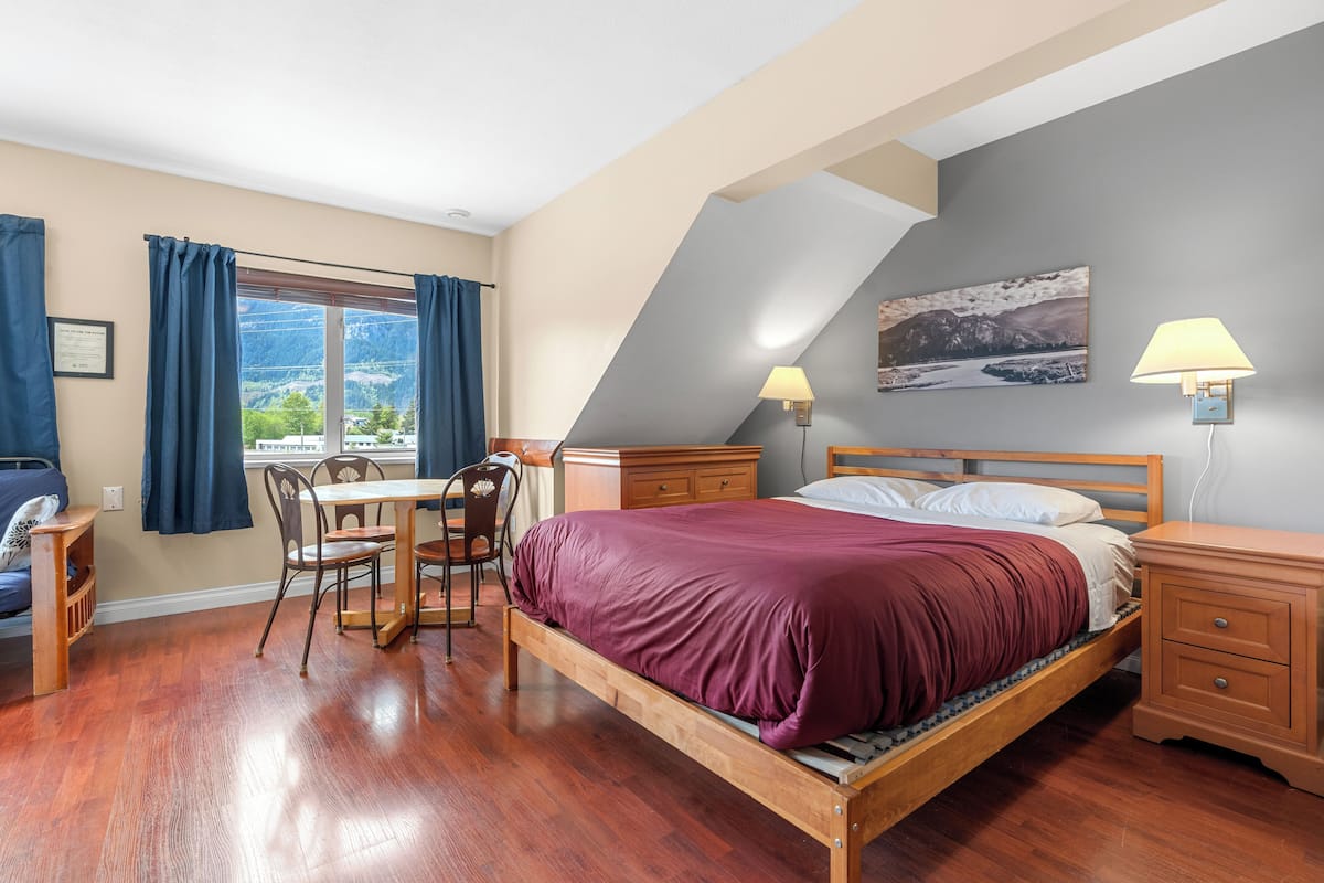 A bedroom with a wooden bed, a desk with a chair, a nightstand, and a window with curtains.