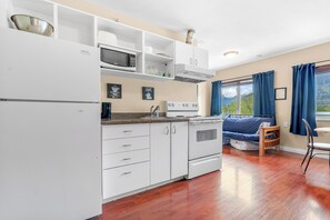 A compact kitchen with white cabinets, a microwave, and a stove. There is a window with blue curtains and a view of greenery outside.
