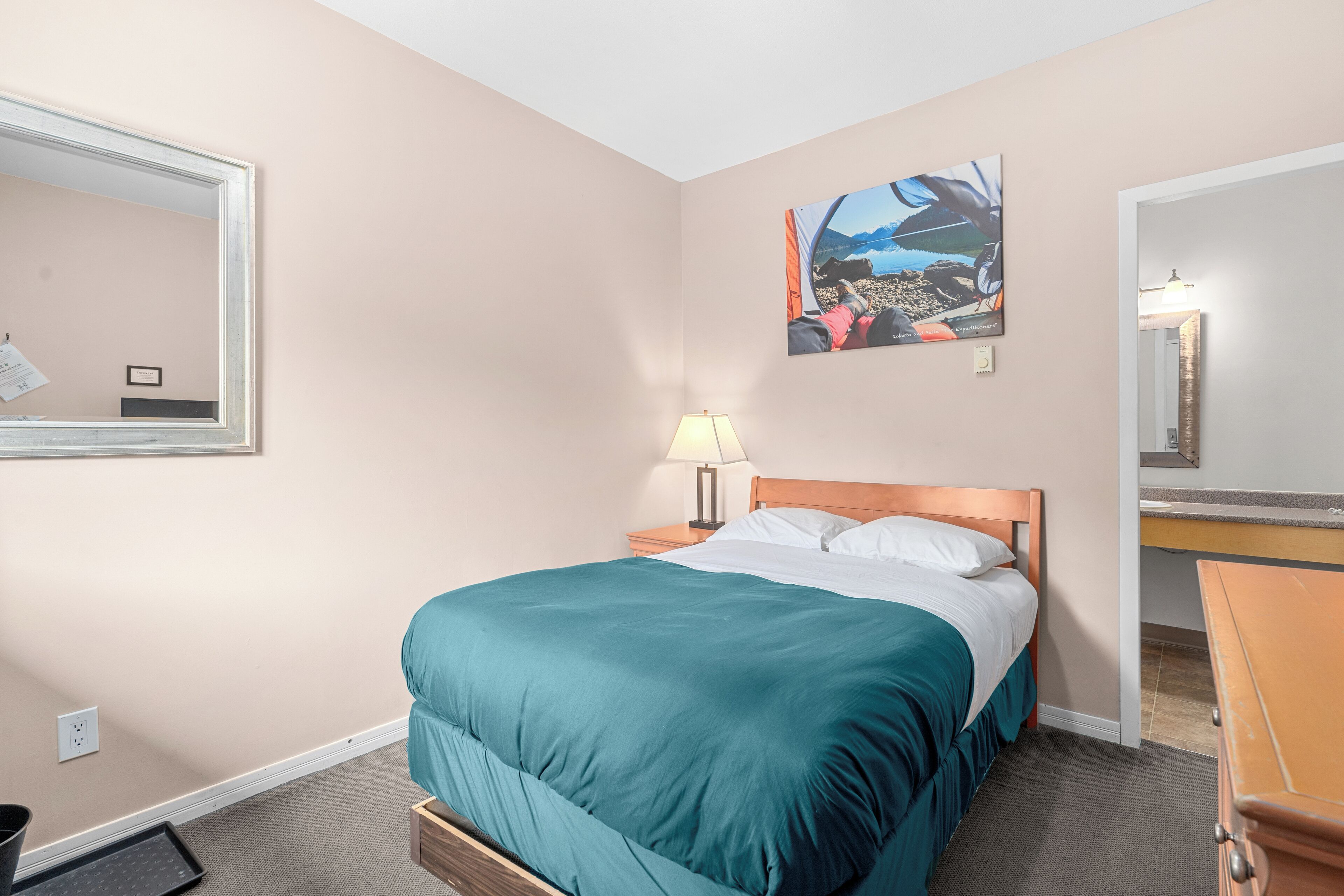 Une chambre d’hôtel avec un lit, une table de chevet, une lampe, une commode et un tableau au mur.