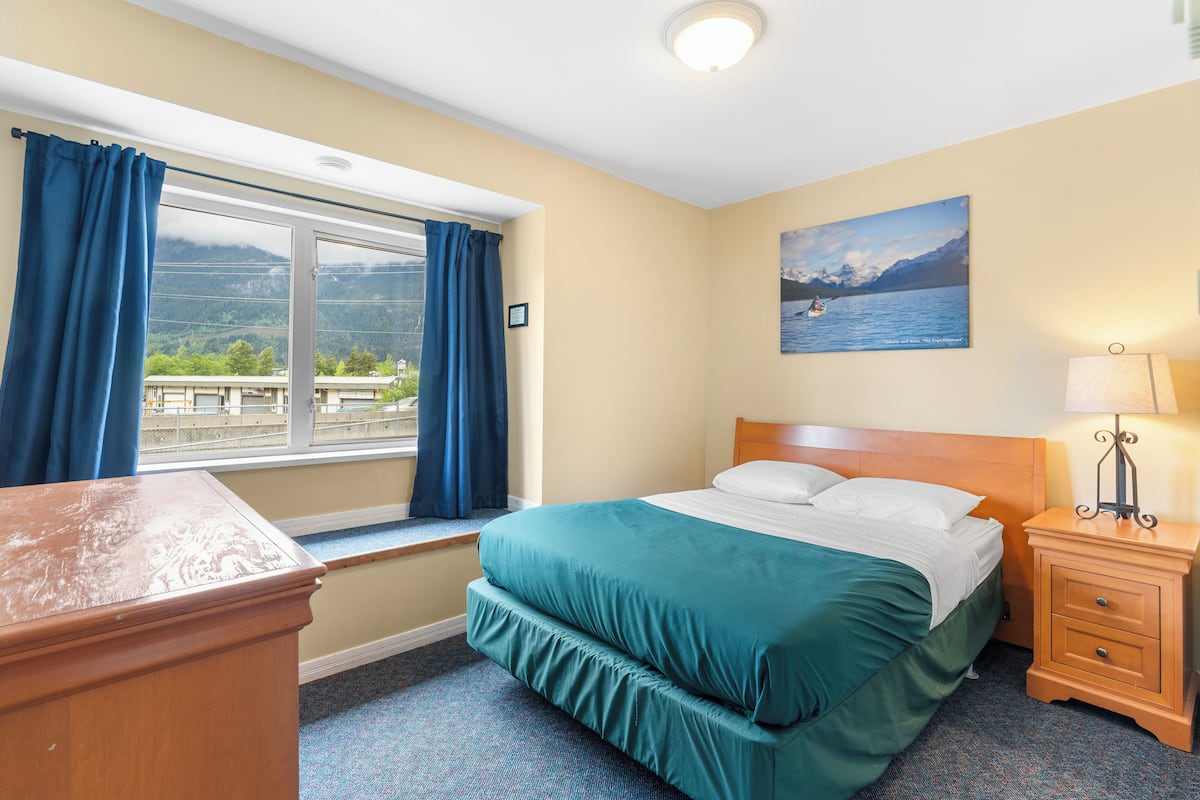 A hotel room with a bed, a desk, a nightstand, a lamp, and a window with curtains.