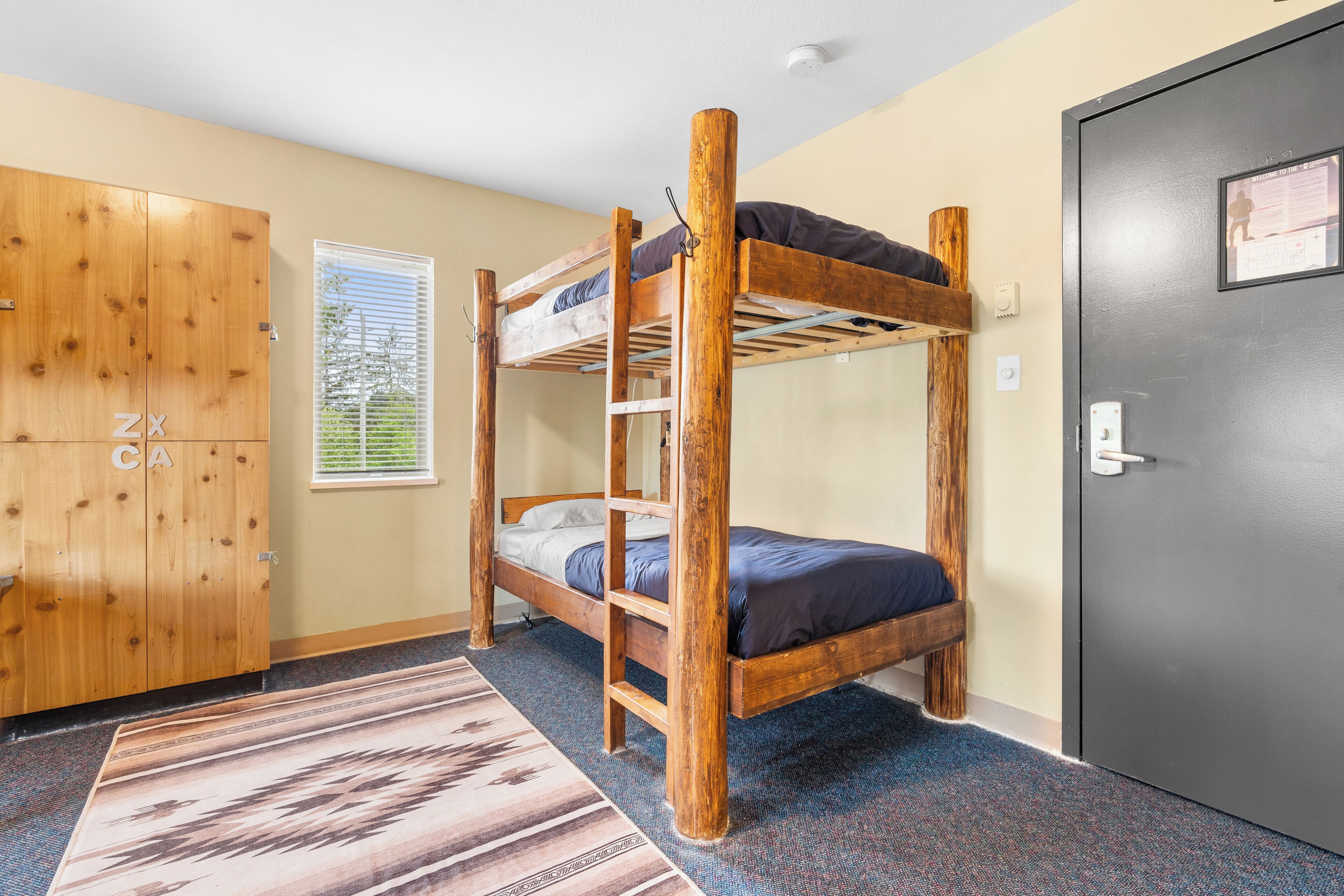 A bunk bed room with a wooden wardrobe, a window with blinds, and a carpeted floor.