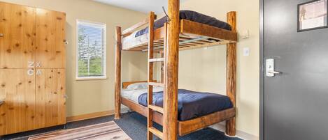 A bunk bed room with a wooden wardrobe, a window with blinds, and a carpeted floor.