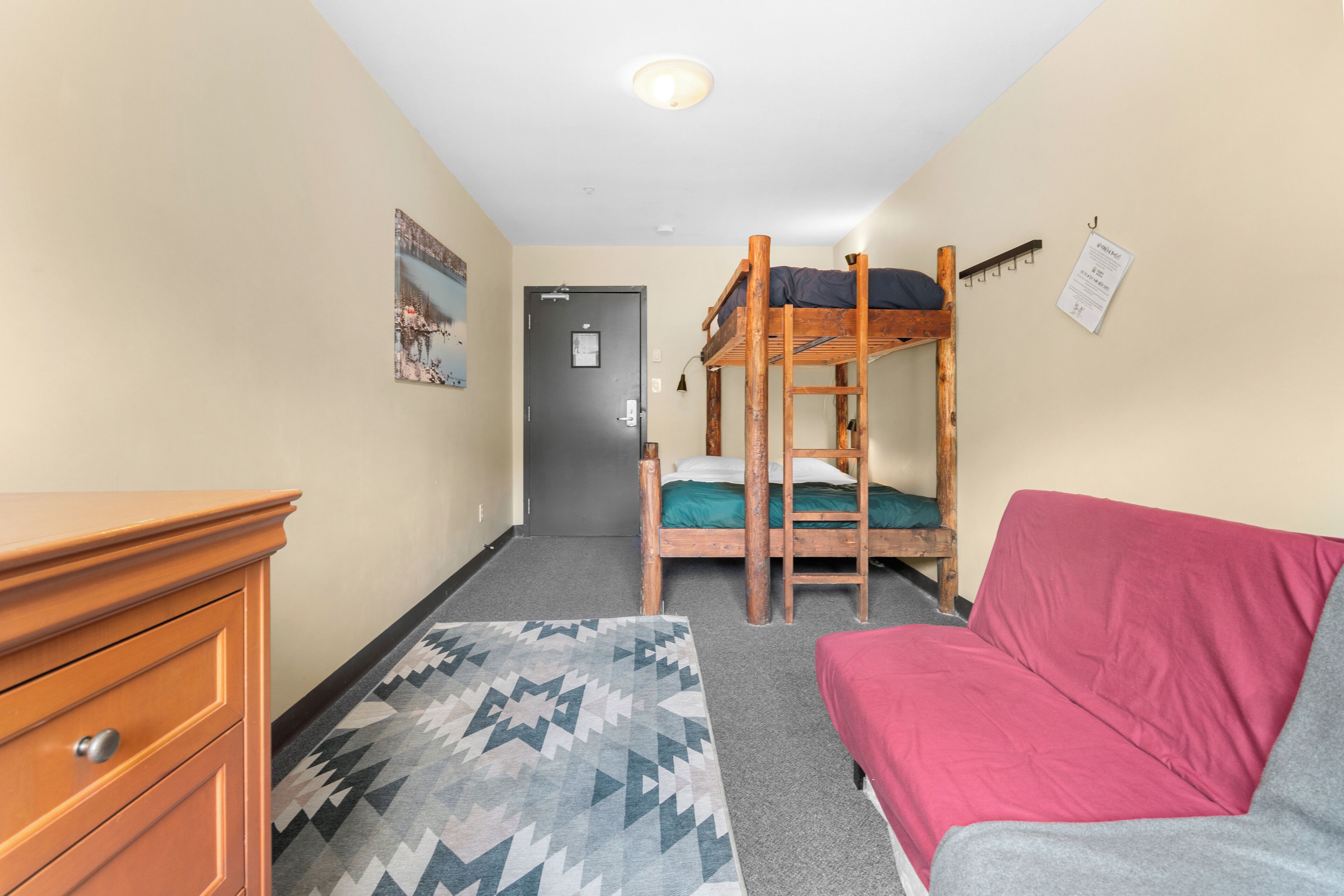A dormitory room with a bunk bed, a wooden dresser, a patterned rug, a red sofa, and a door with a notice on the wall.