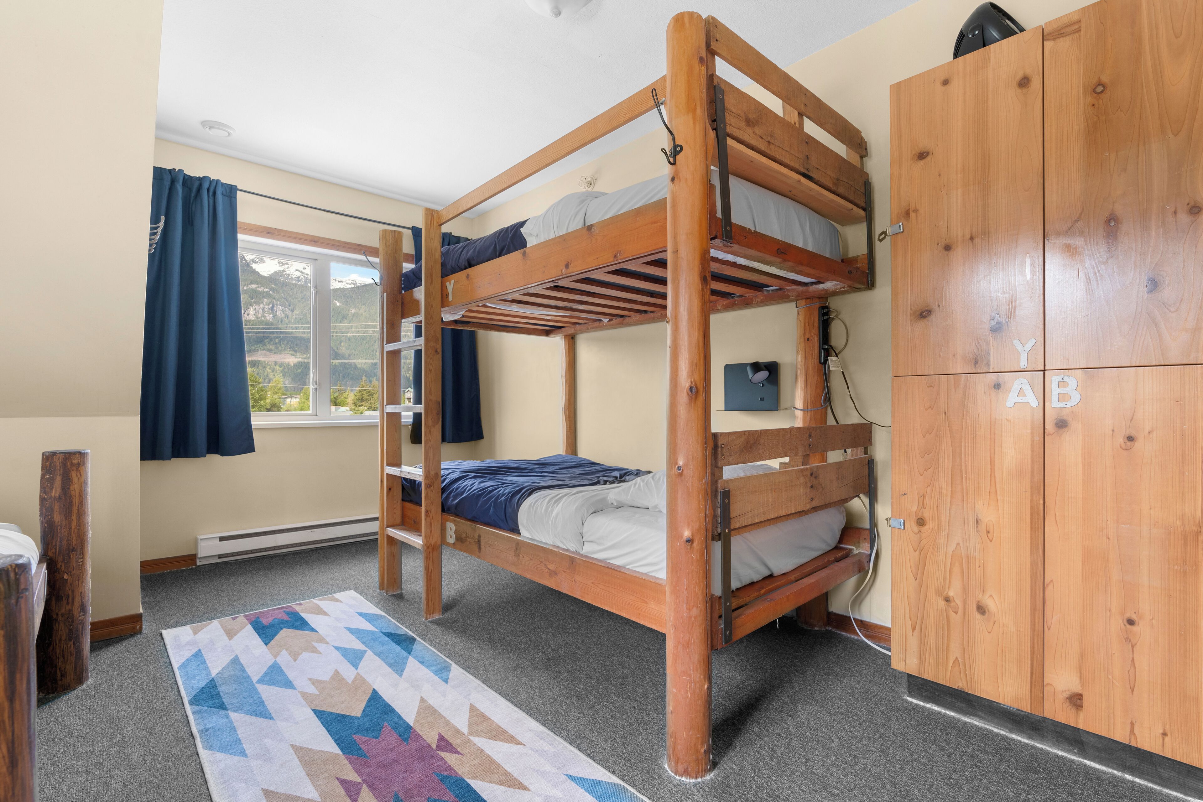A bunk bed room with a wooden wardrobe, a window with curtains, and a patterned rug.