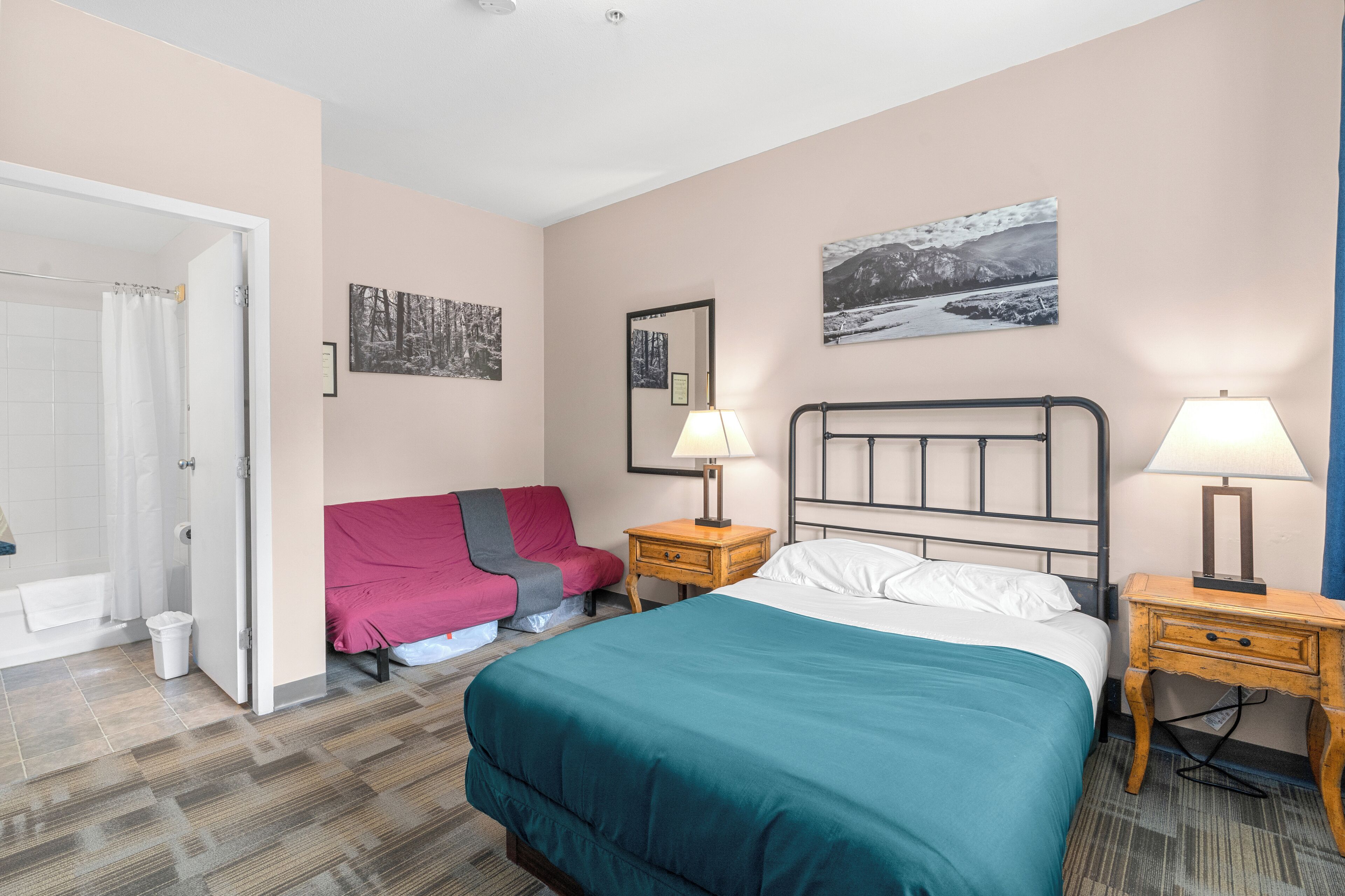 Habitación de hotel con cama, dos mesitas de noche con lámparas, una silla roja y zona de ducha.