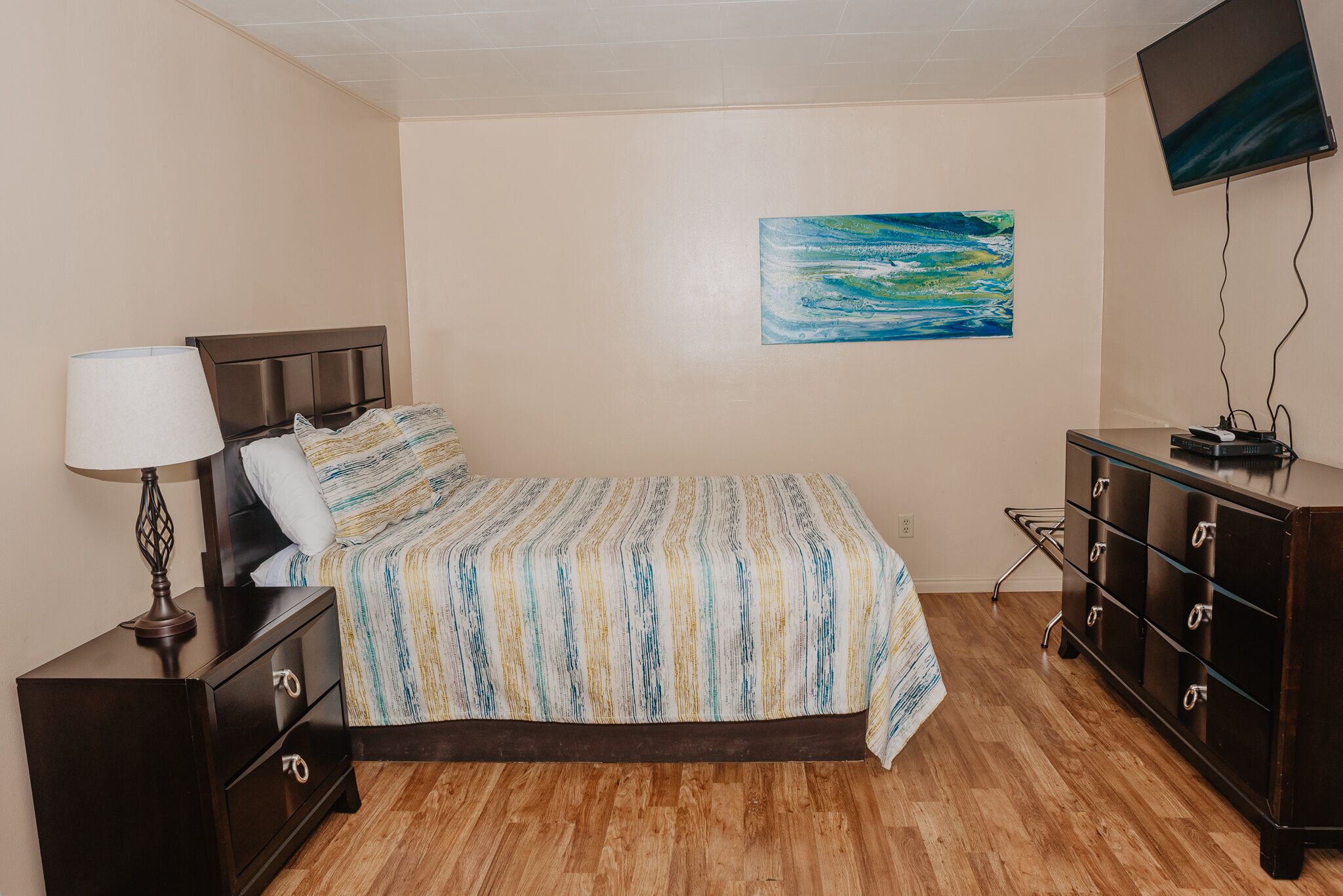 Une chambre à coucher comprenant un lit, une table de chevet, une commode, un tableau accroché au mur et un téléviseur fixé au mur.