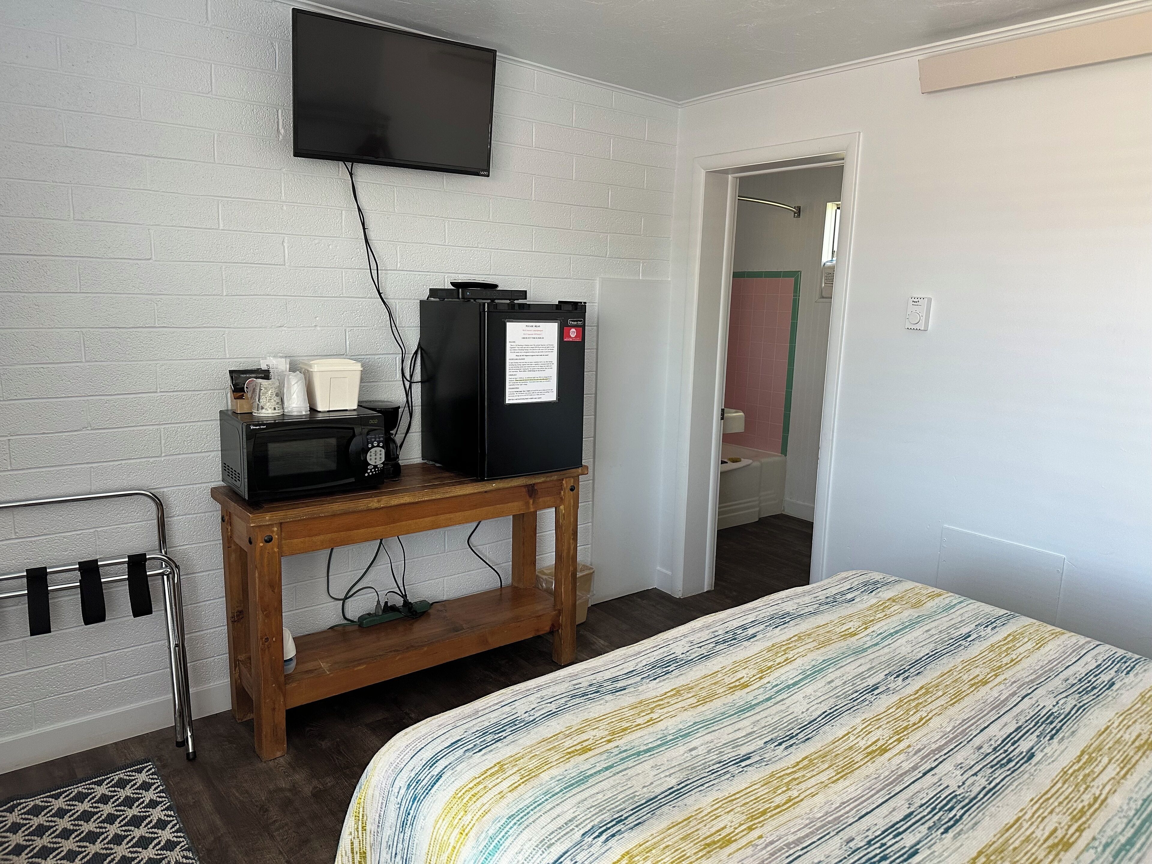 A room with a bed, a TV mounted on the wall, a microwave, a refrigerator, and a wooden table with a coffee maker and cups.