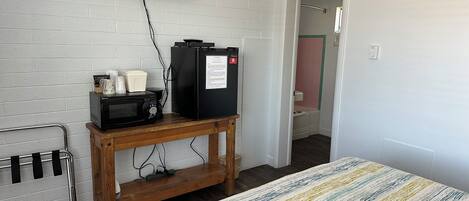 A room with a bed, a TV mounted on the wall, a microwave, a refrigerator, and a wooden table with a coffee maker and cups.