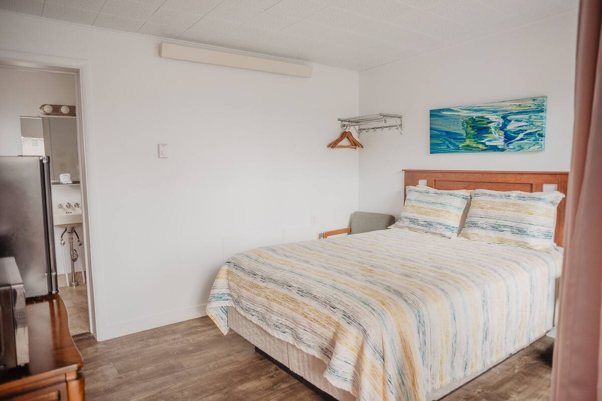 A bedroom with a bed, a wooden headboard, a painting on the wall, and a hallway leading to a bathroom.
