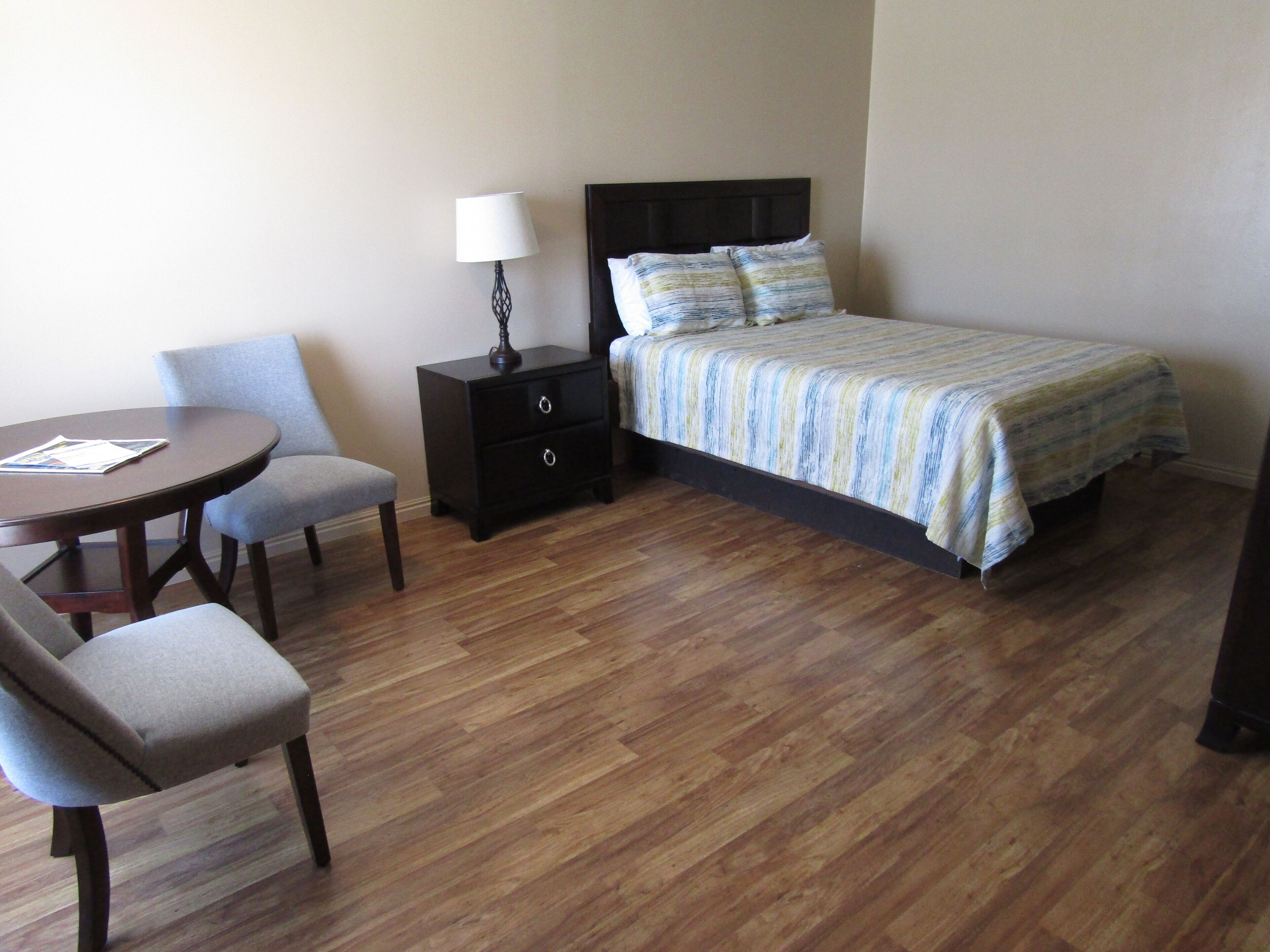 A hotel room with a bed, a nightstand with a lamp, a small table with two chairs, and wooden flooring.