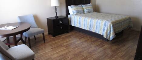 A hotel room with a bed, a nightstand with a lamp, a small table with two chairs, and wooden flooring.