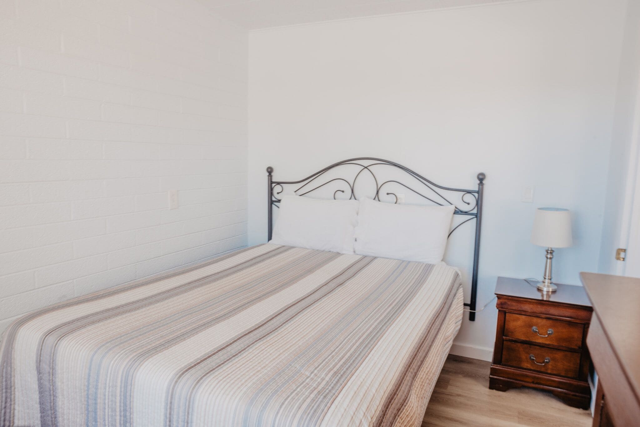 Un lit simple avec une couvre-lit à rayures, une table de chevet en bois avec une lampe et un mur en briques blanches.