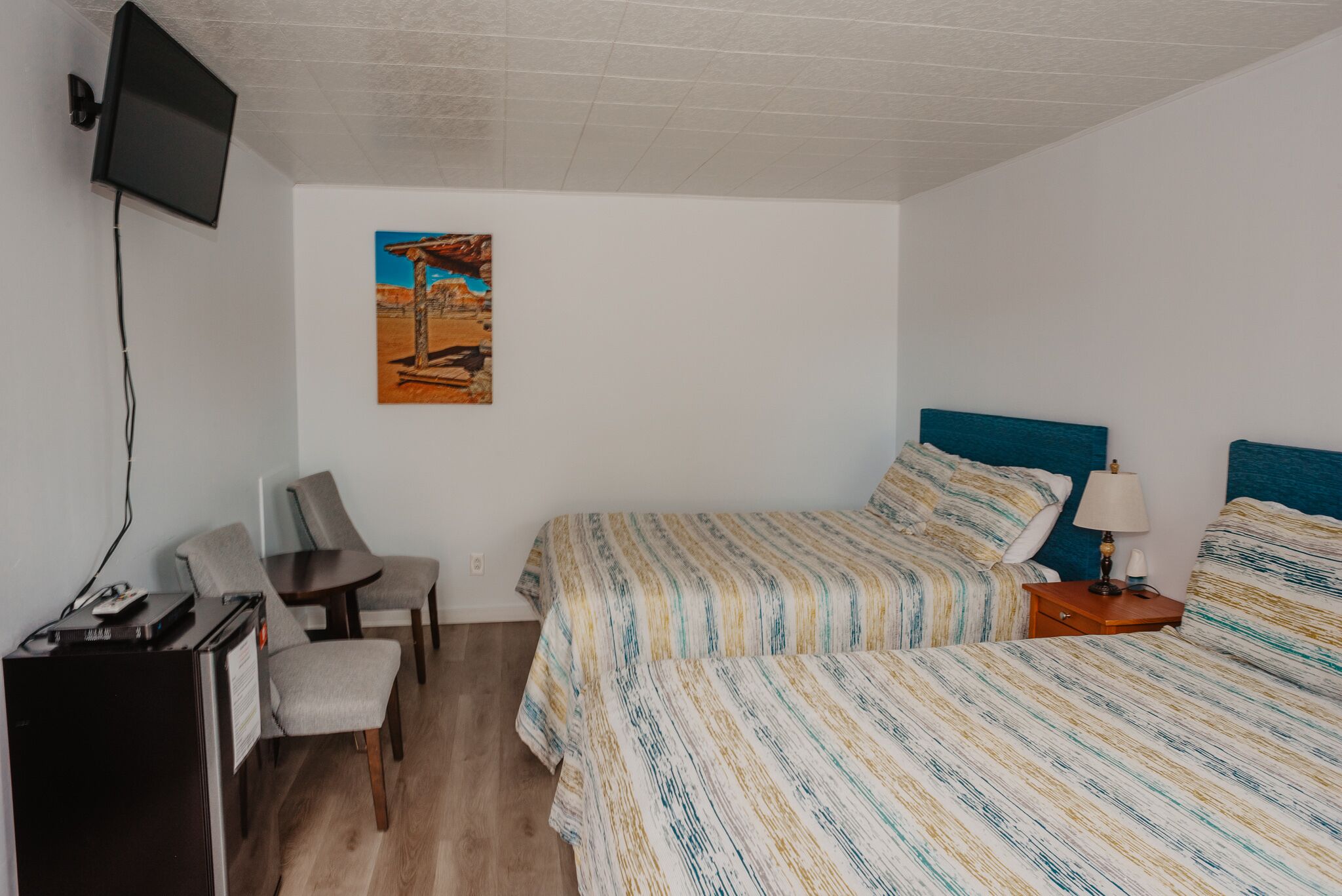 A bedroom with two beds, a desk, a chair, a TV mounted on the wall, and a painting.