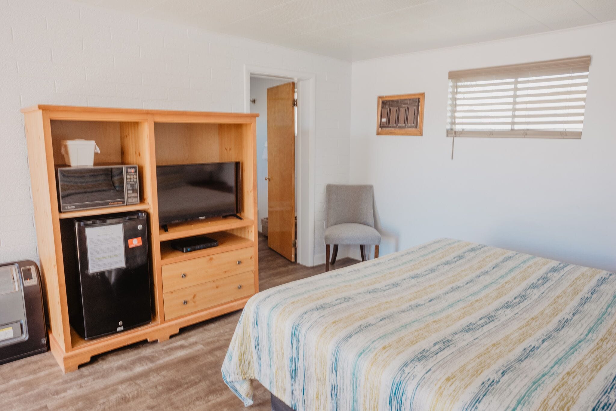 A bedroom with a bed, a wooden TV stand with a microwave and a mini-fridge, a grey chair, and a window with blinds.