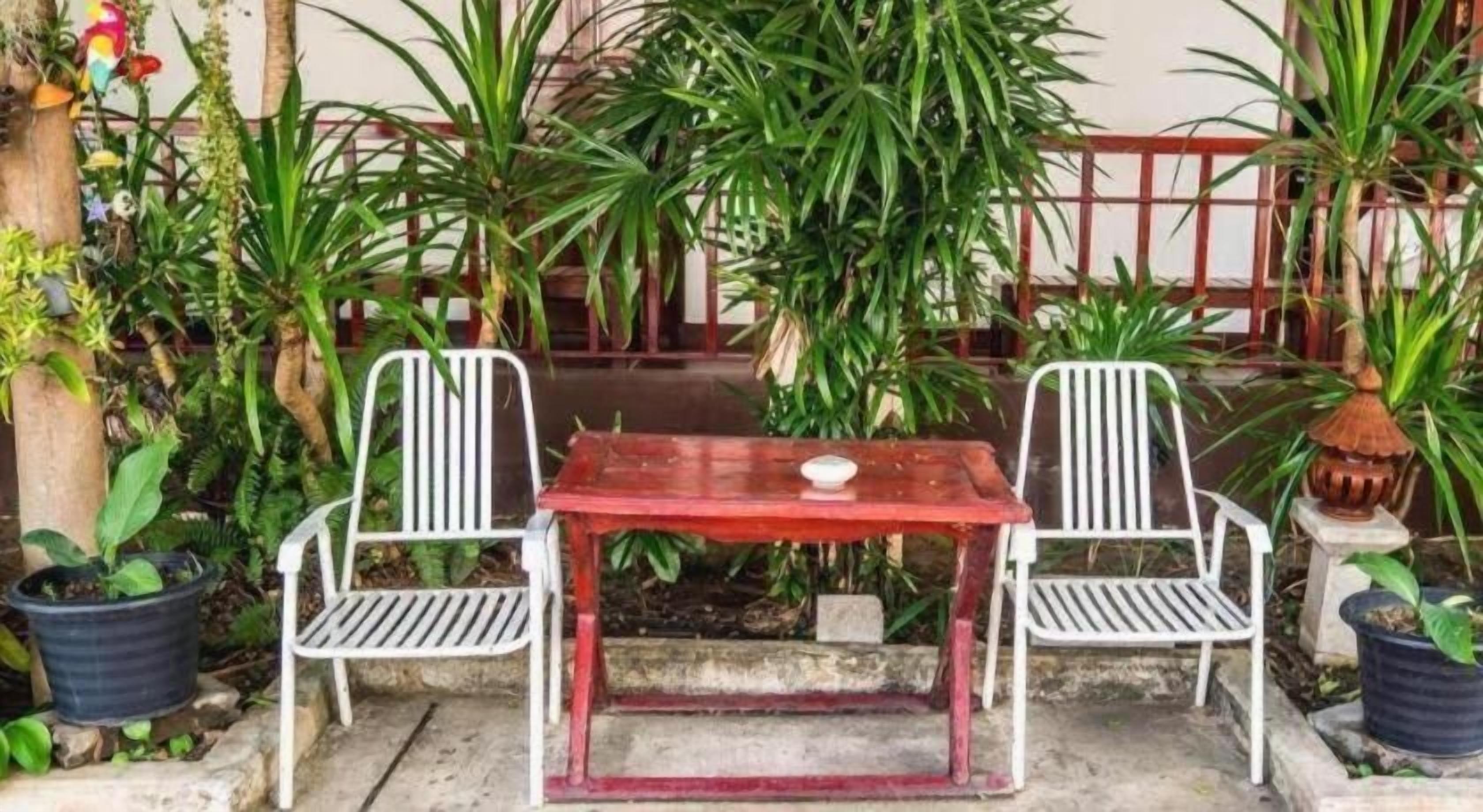 Un espacio exterior con asientos, dos sillas de metal blanco y una mesa roja, rodeado de plantas en macetas y una cerca de metal.