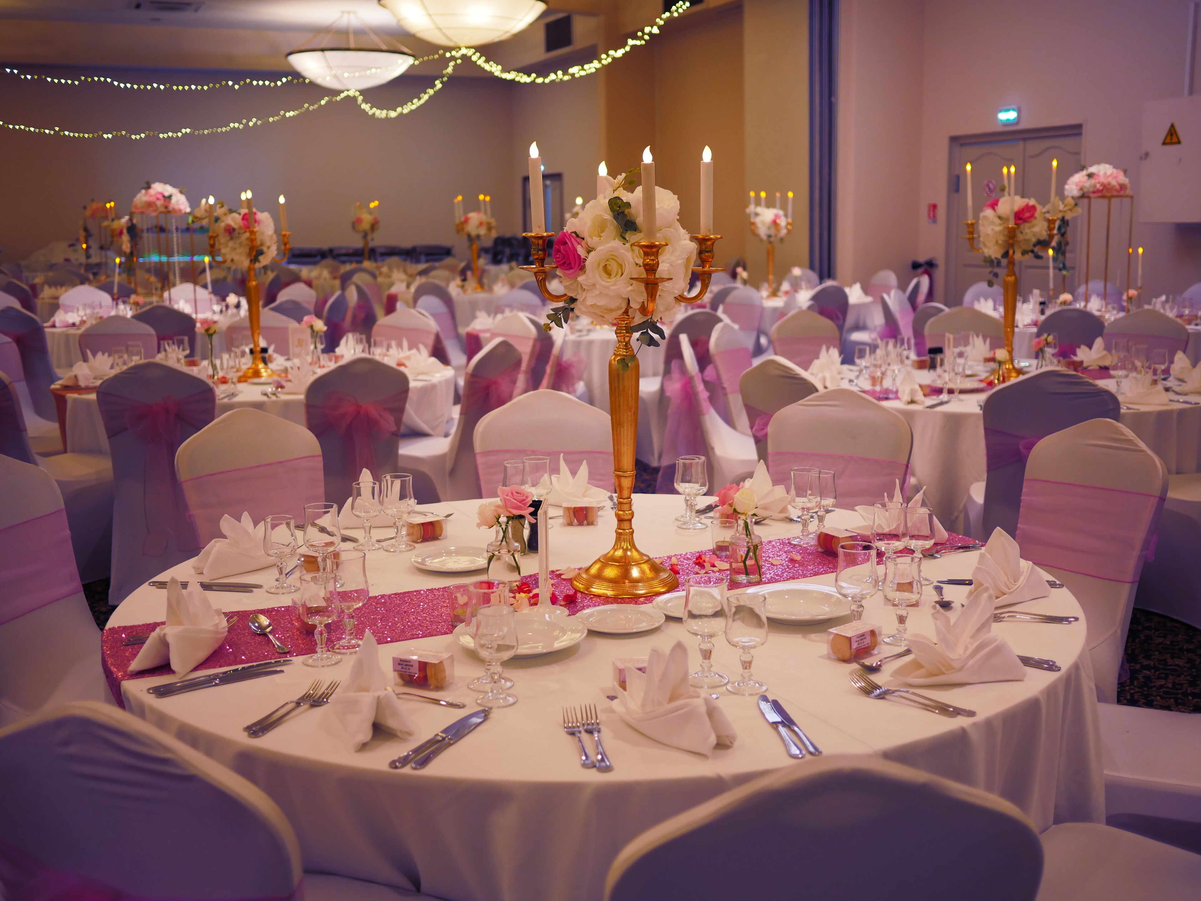 Un salón de banquetes con mesas redondas dispuestas para un evento formal, cada una con manteles blancos, cintas rosas y centros de mesa florales.