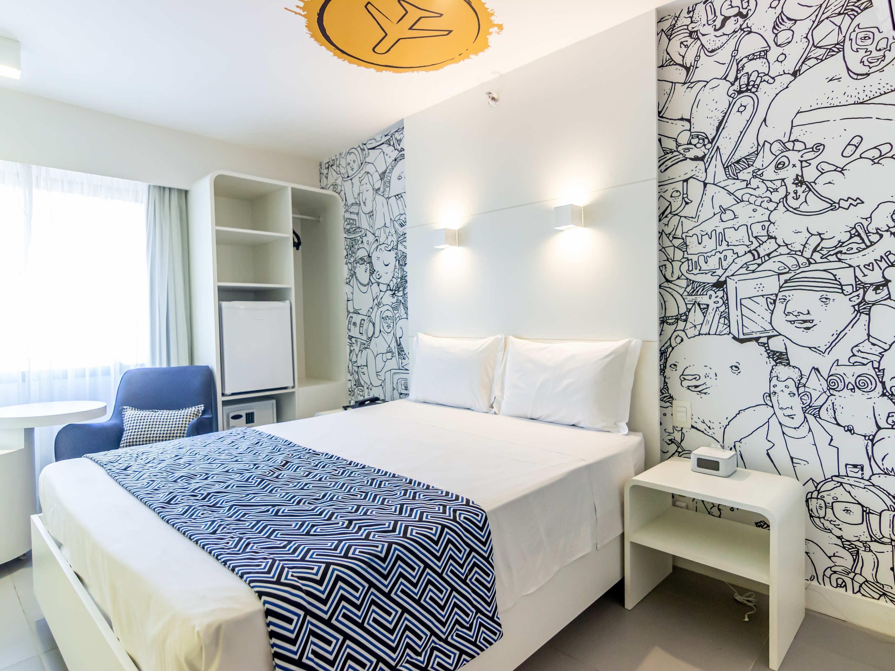 A hotel room with a bed, a desk, a chair, a wardrobe, and a wall with a black and white mural.