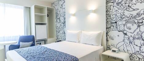 A hotel room with a bed, a desk, a chair, a wardrobe, and a wall with a black and white mural.