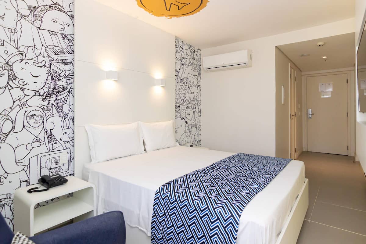 A hotel room with a bed, bedside table, and a chair. There is a wall with a black and white mural.